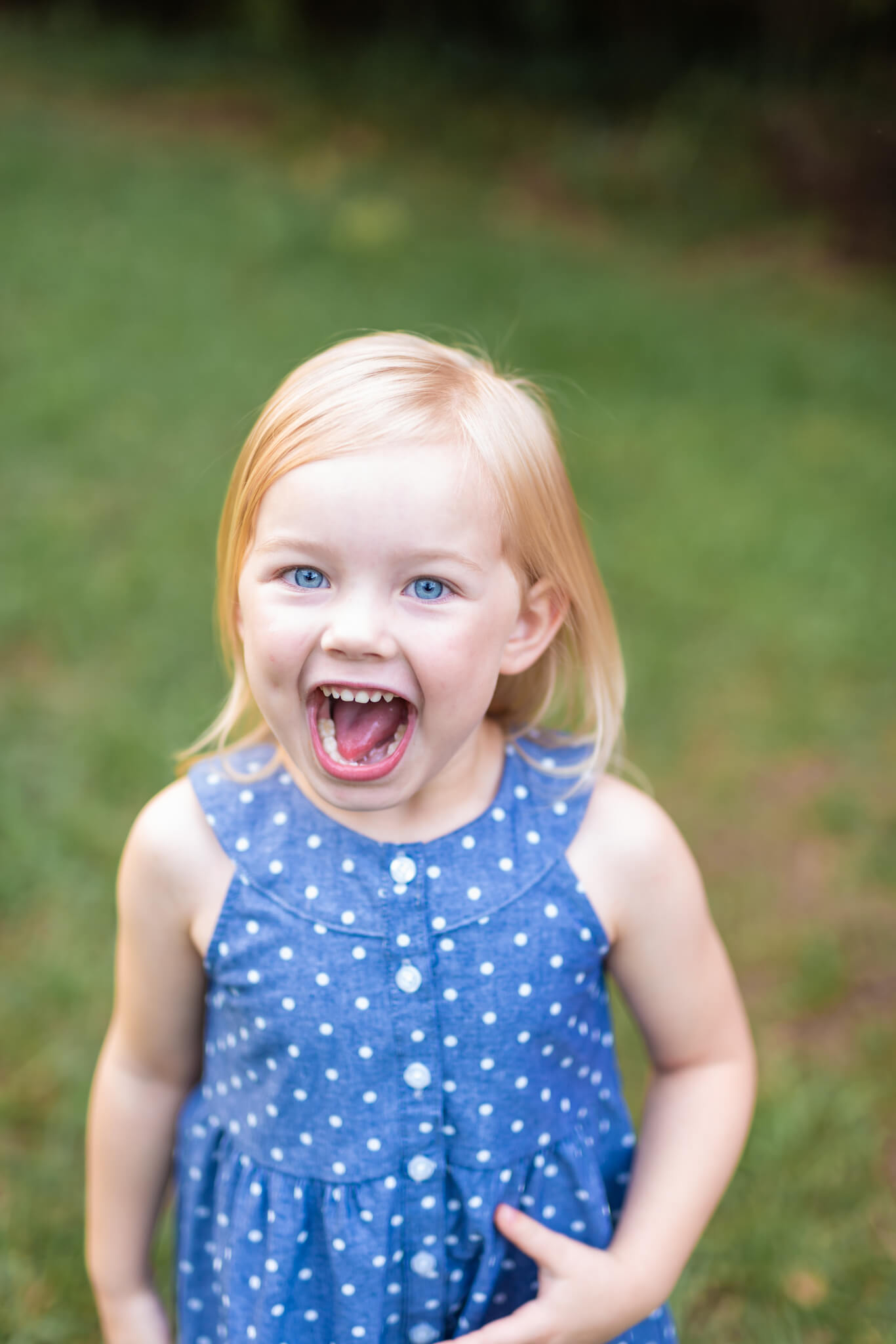An excited toddler girl in a blue polka dot dress stands in a park lawn