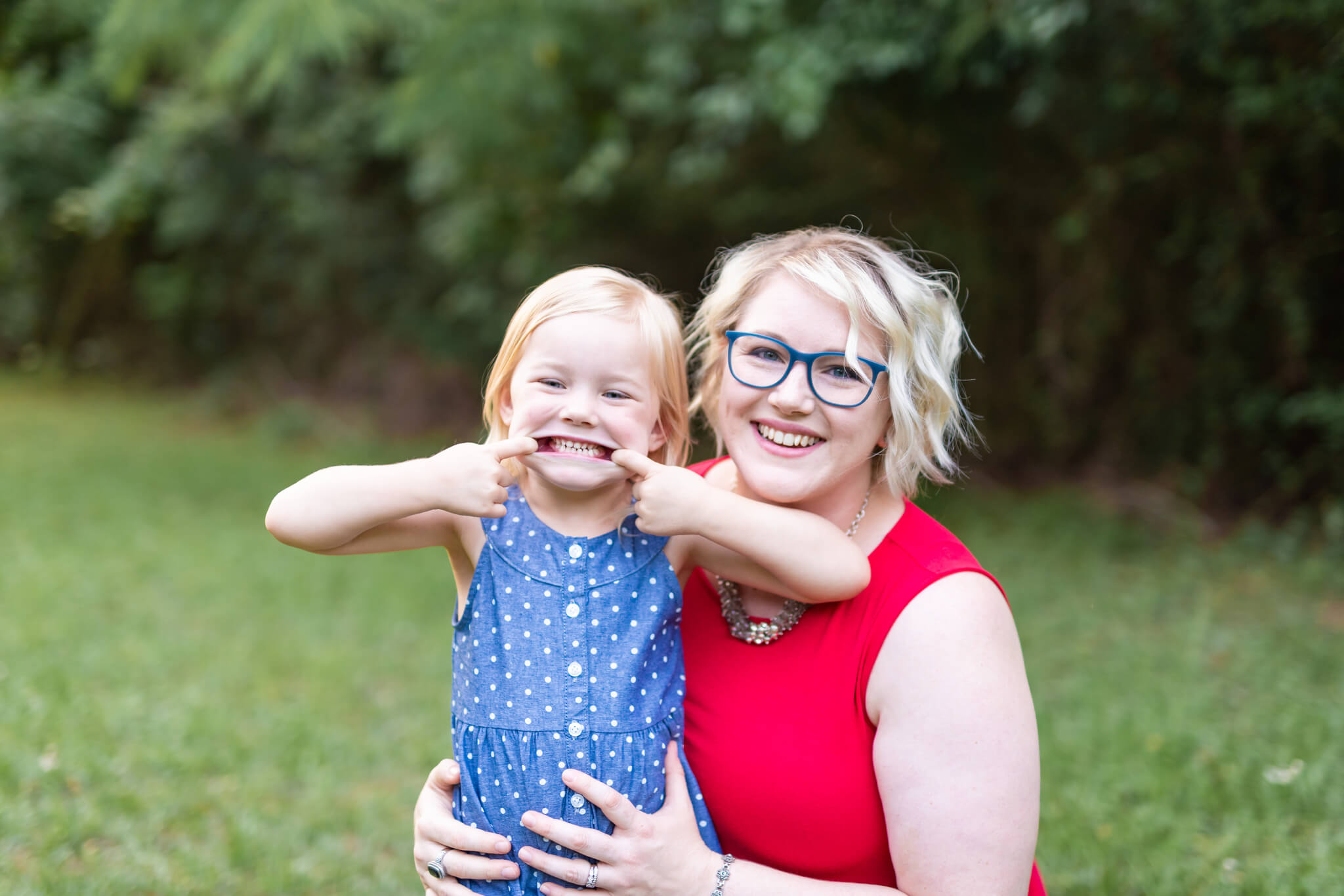 A toddler girl in a blue dress makes a silly face while standing with her mom in a park in red after some swim lessons in savannah ga