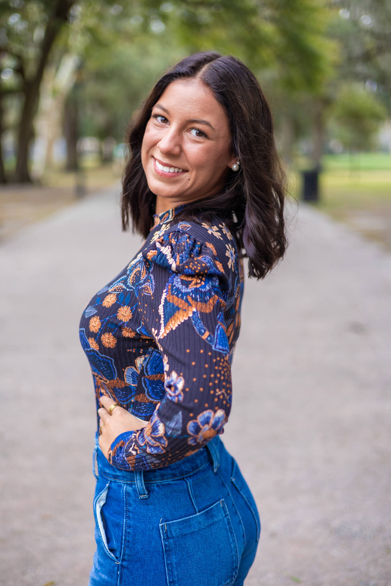 A smiling woman in a blue flower print blouse stands smiling over her shoulder in a park sidewalk
