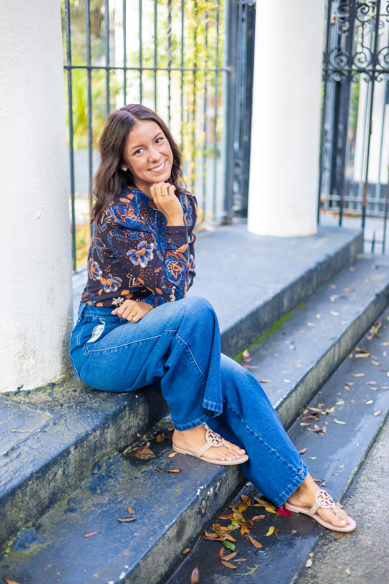 A smiling high school senior in jeans and flower print blouse sits on park steps smiling after visiting tanning salons in savannah ga