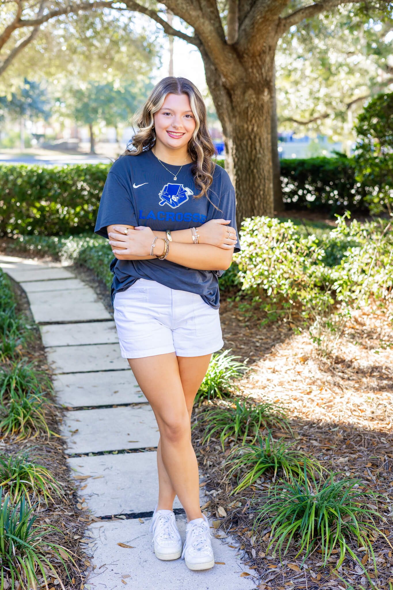 A high school senior stands in a garden path with arms crossed in a blue lacrosse shirt and white shorts with a big smile