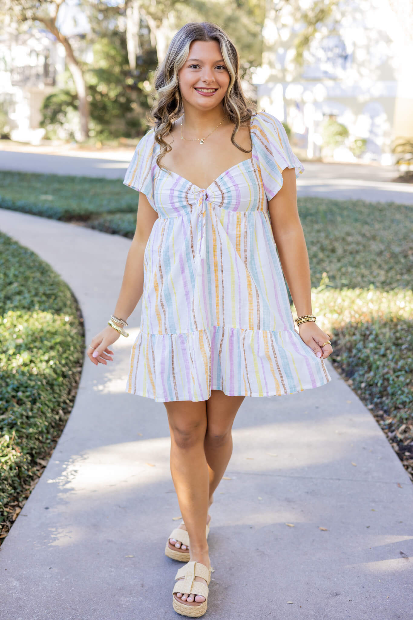 A high school senior in a colorful dress walks in a garden sidewalk smiling