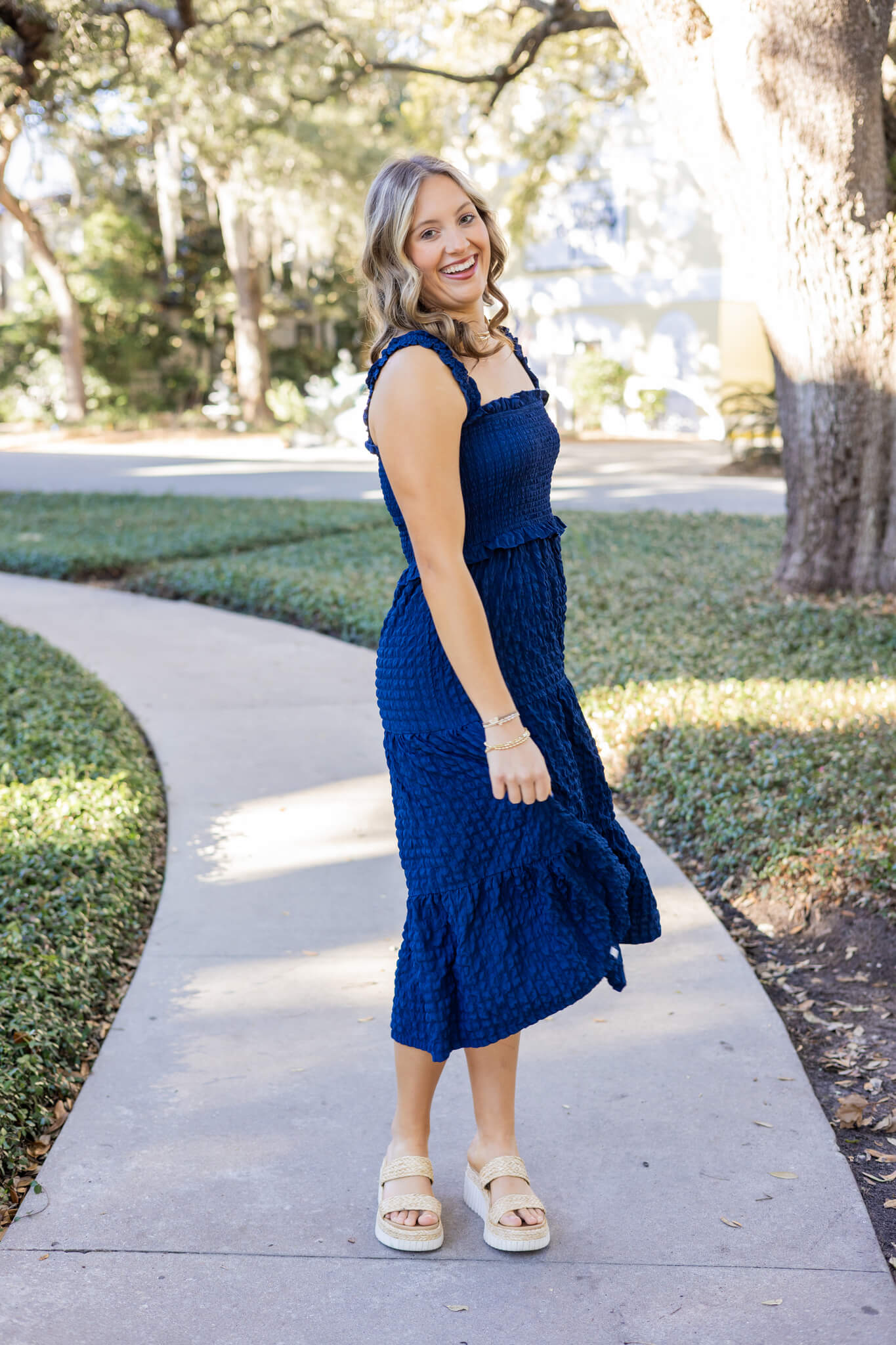 A woman in a blue dress smiles while standing on a sidewalk in a park and twirling after shopping boutiques in pooler ga