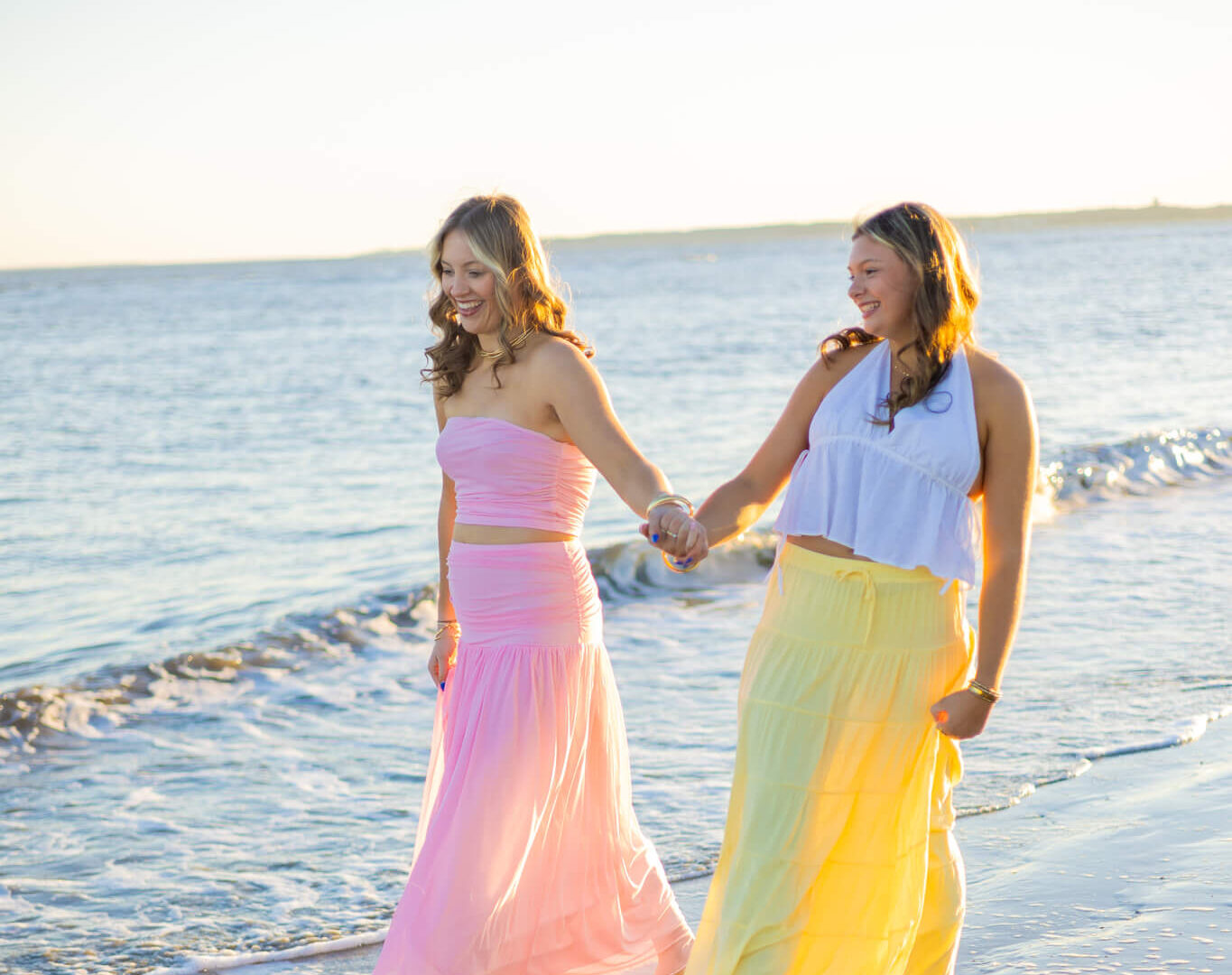 High school senior girls walk holding hands on a beach in bright colored long skirts at sunset after finding great boutiques in pooler ga