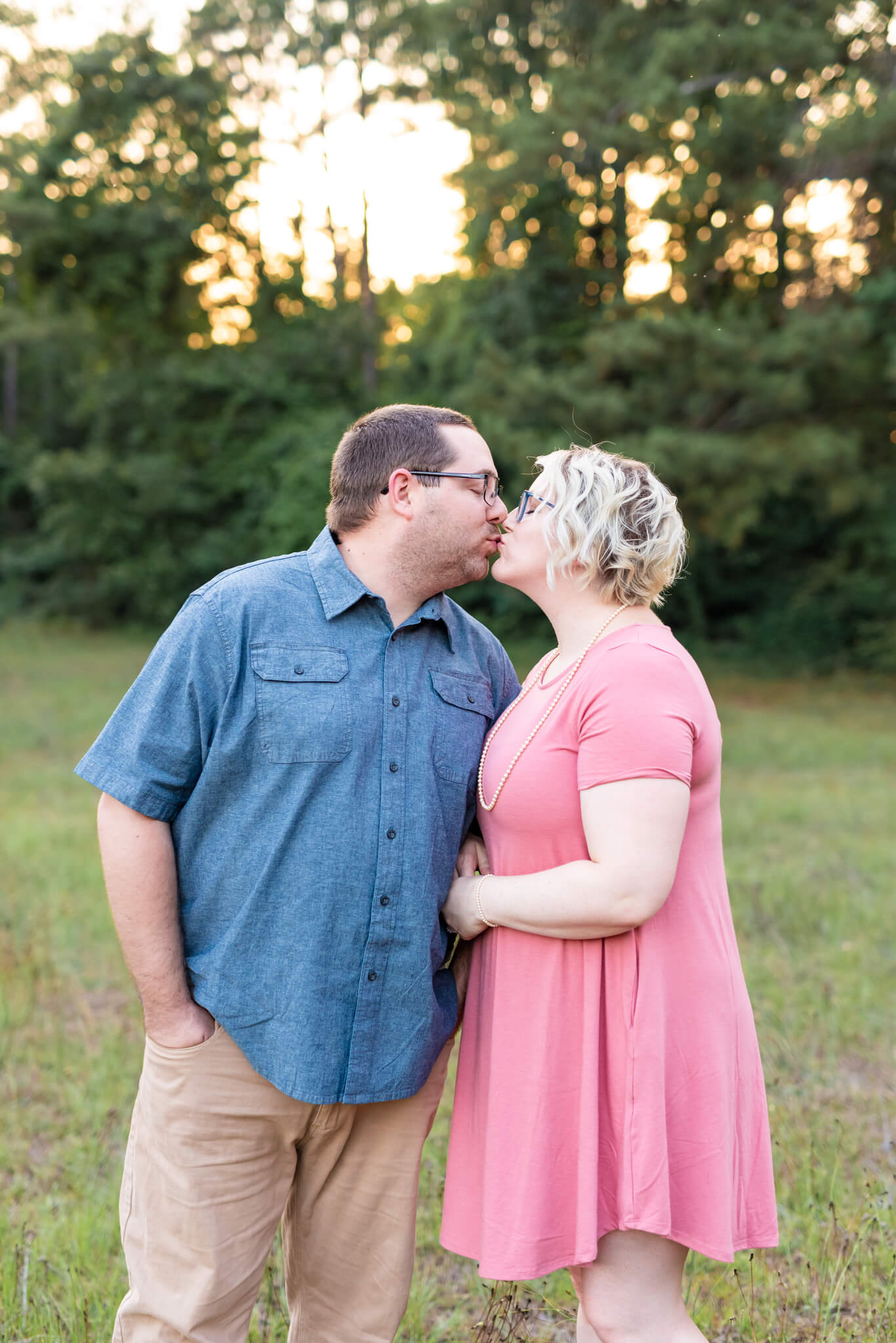 A happy couple kisses in a park in a pink dress and blue shirt with hands in pockets