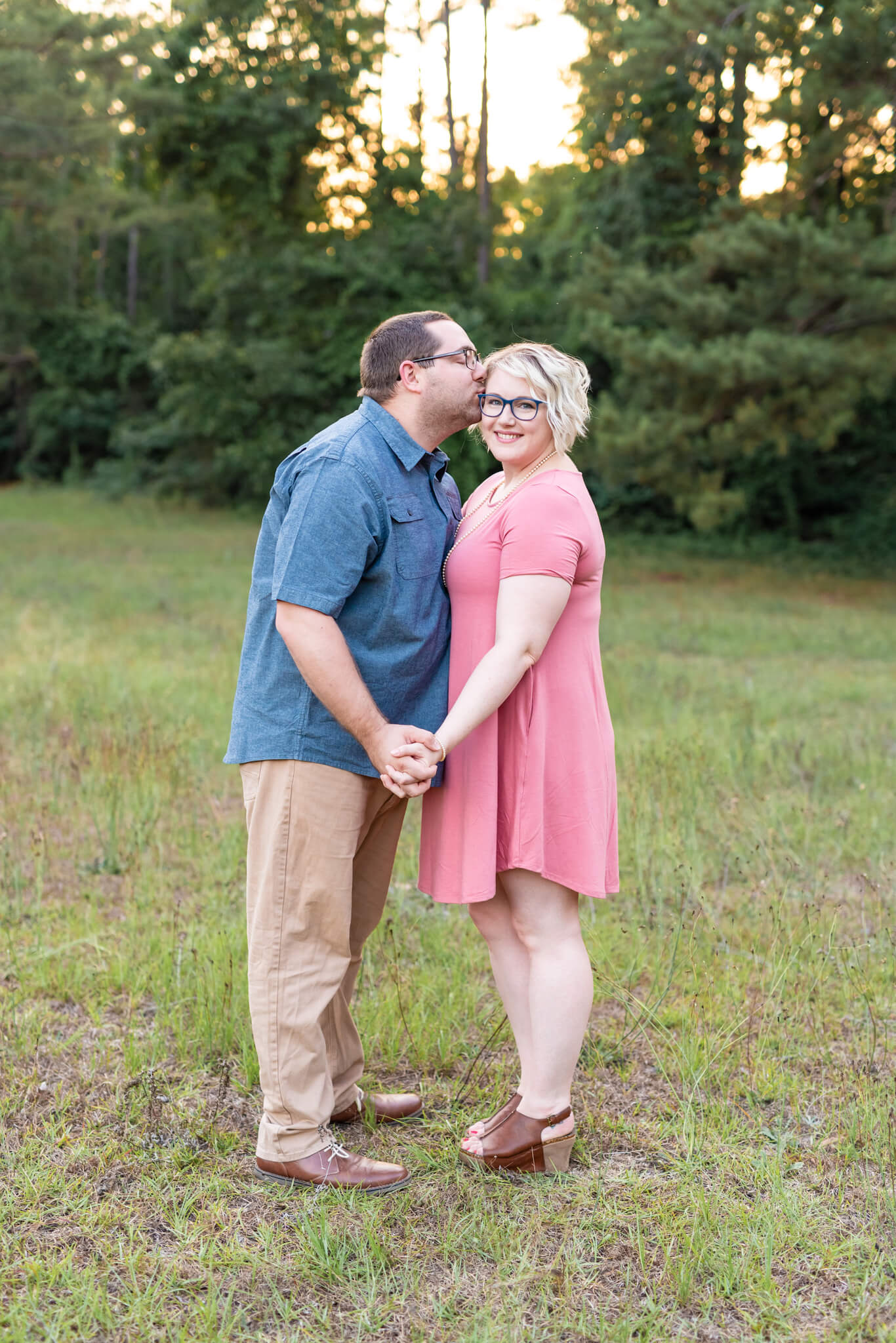 A happy woman in a pink dress is kissed on the cheek by her husband in blue in a park lawn at sunset after some couples cooking classes in savannah, ga