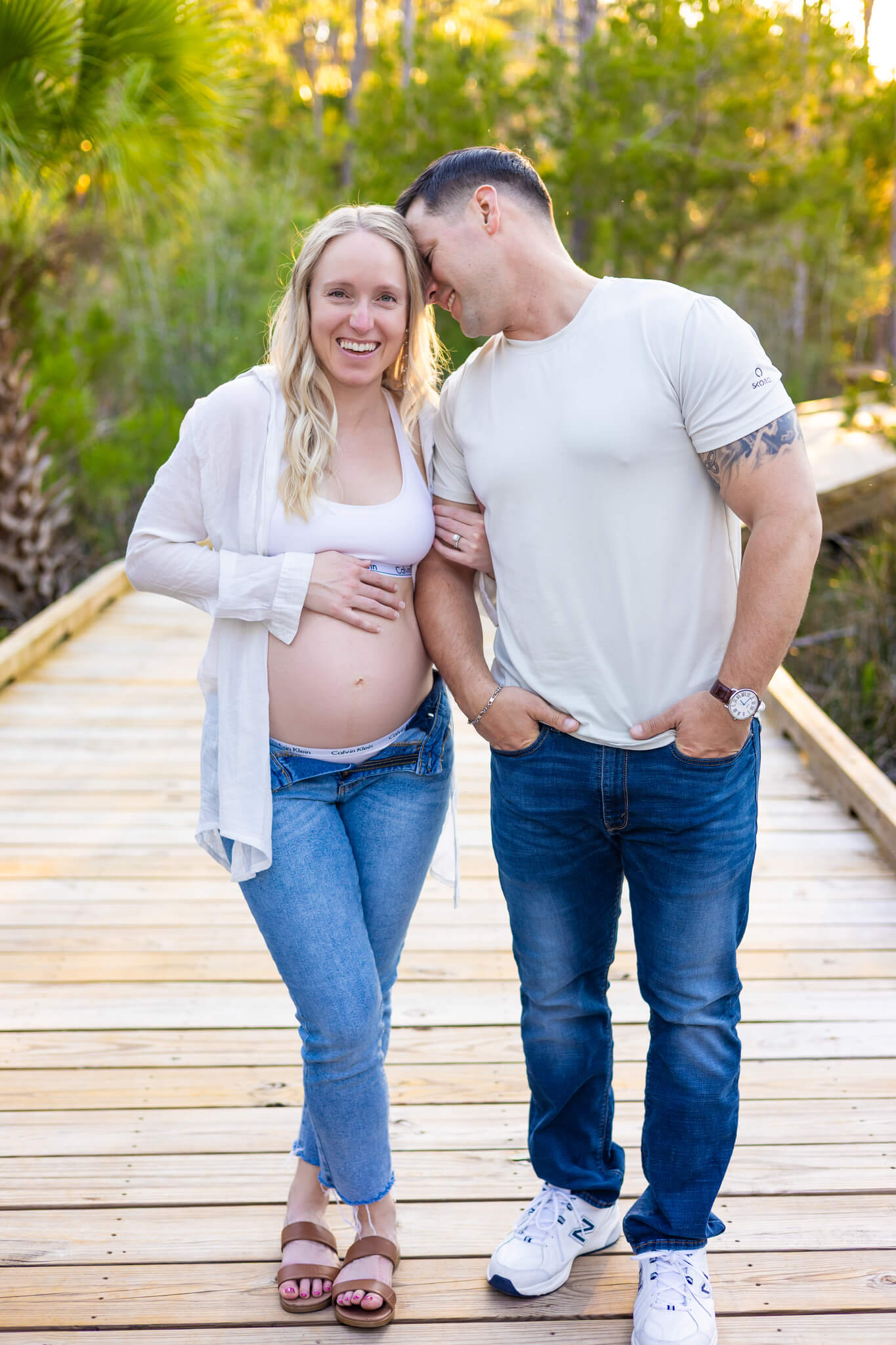 A pregnant woman laughs while walking on a boardwalk at sunset arm in arm with her snuggling husband