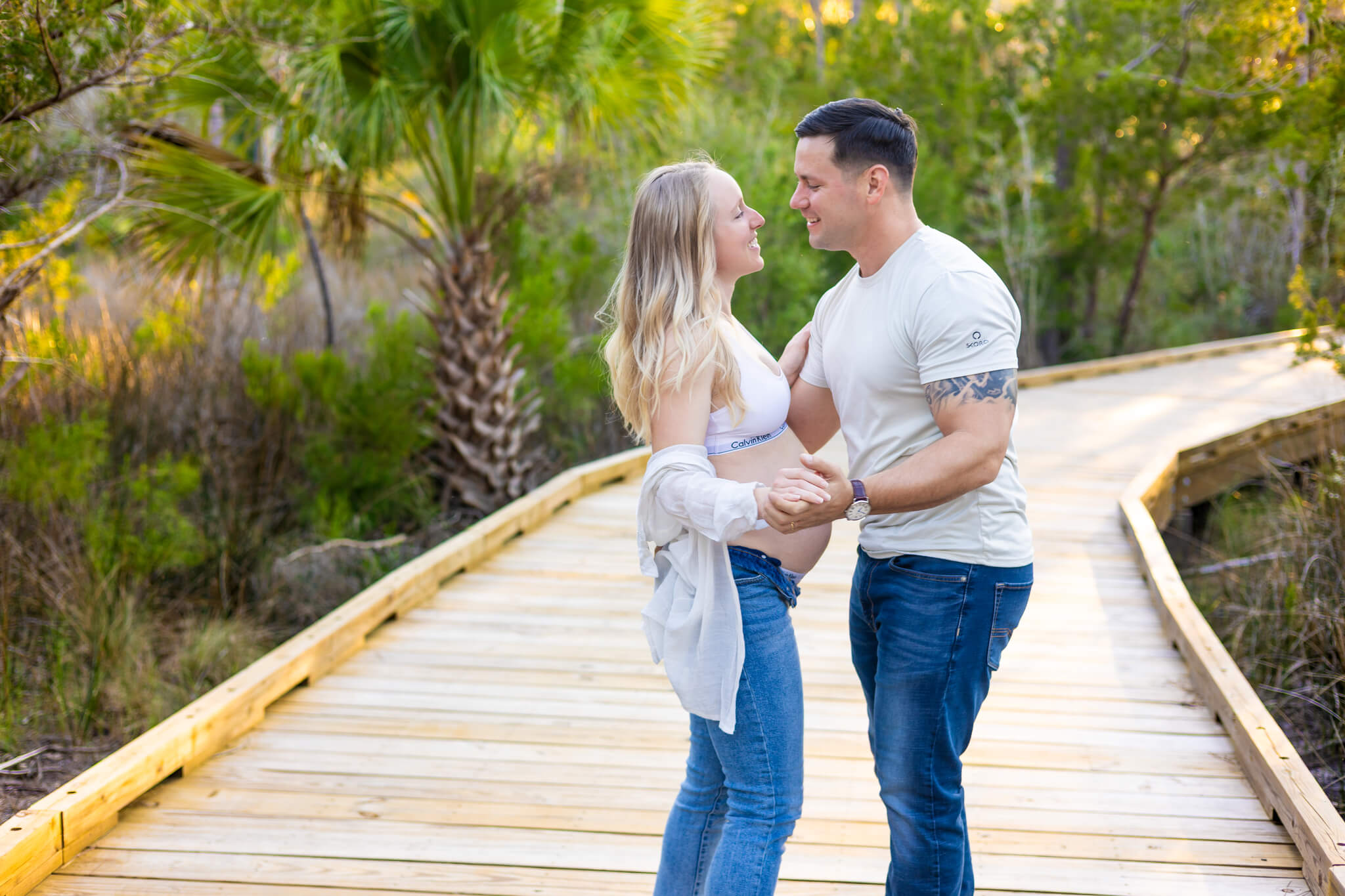 A happy pregnant couple dances on a boardwalk through a wetland at sunset in jeans and white shirts after enjoying day spas in savannah ga