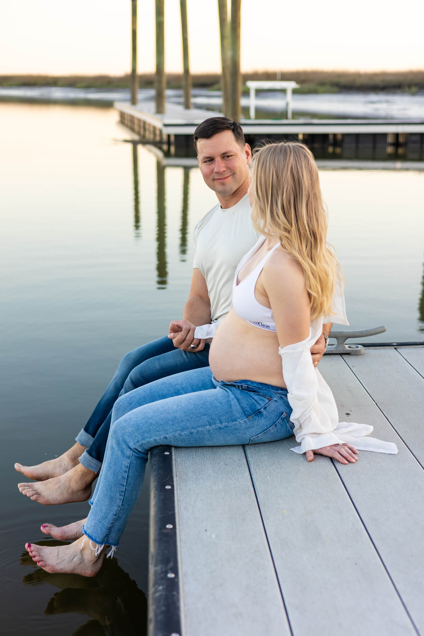 A happy pregnant couple in jeans and white tops sit on the edge of a dock with feet over the water after finding day spas in savannah ga