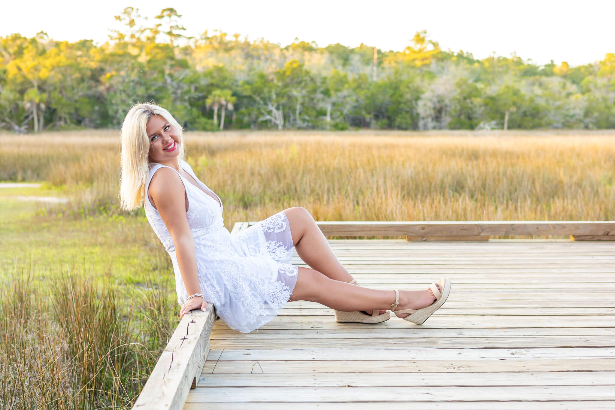 A smiling woman sits on a wooden dock edge in a white lace dress overlooking wetlands at sunset