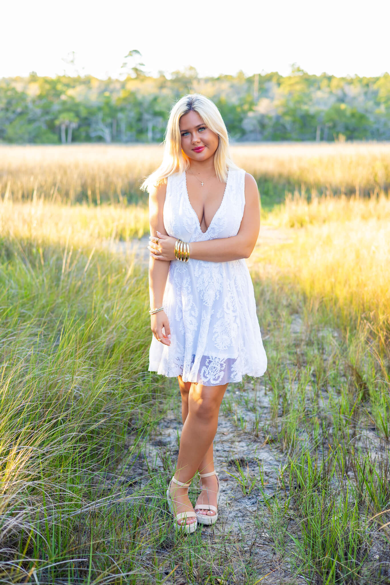 A high school senior in a white lace gown stands in a trail at sunset holding one arm after finding event spaces in savannah, ga