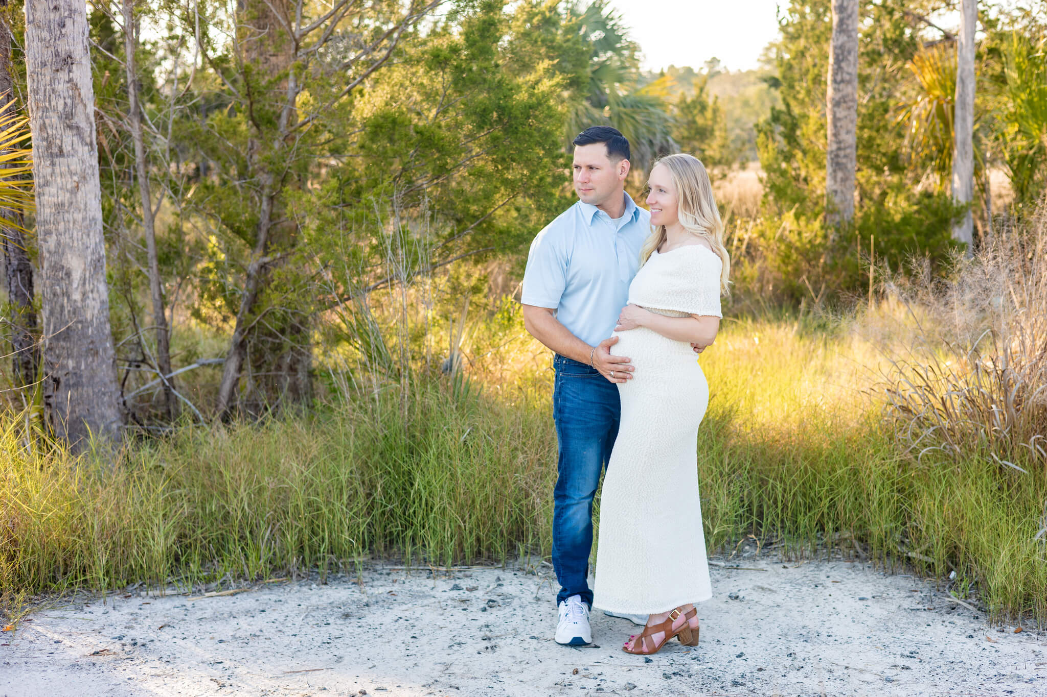 An expectant couple stands admiring the sunset in a Savannah park trail in yellow and blue outfits