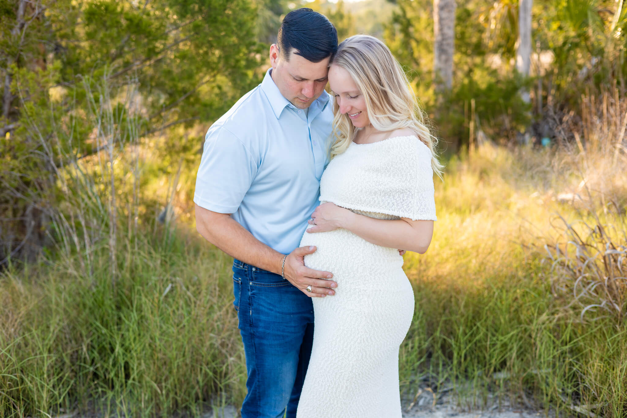 A pregnant couple stands with hands on the bump in blue and yellow in a park filled with tall grasses after finding places to propose in savannah, ga