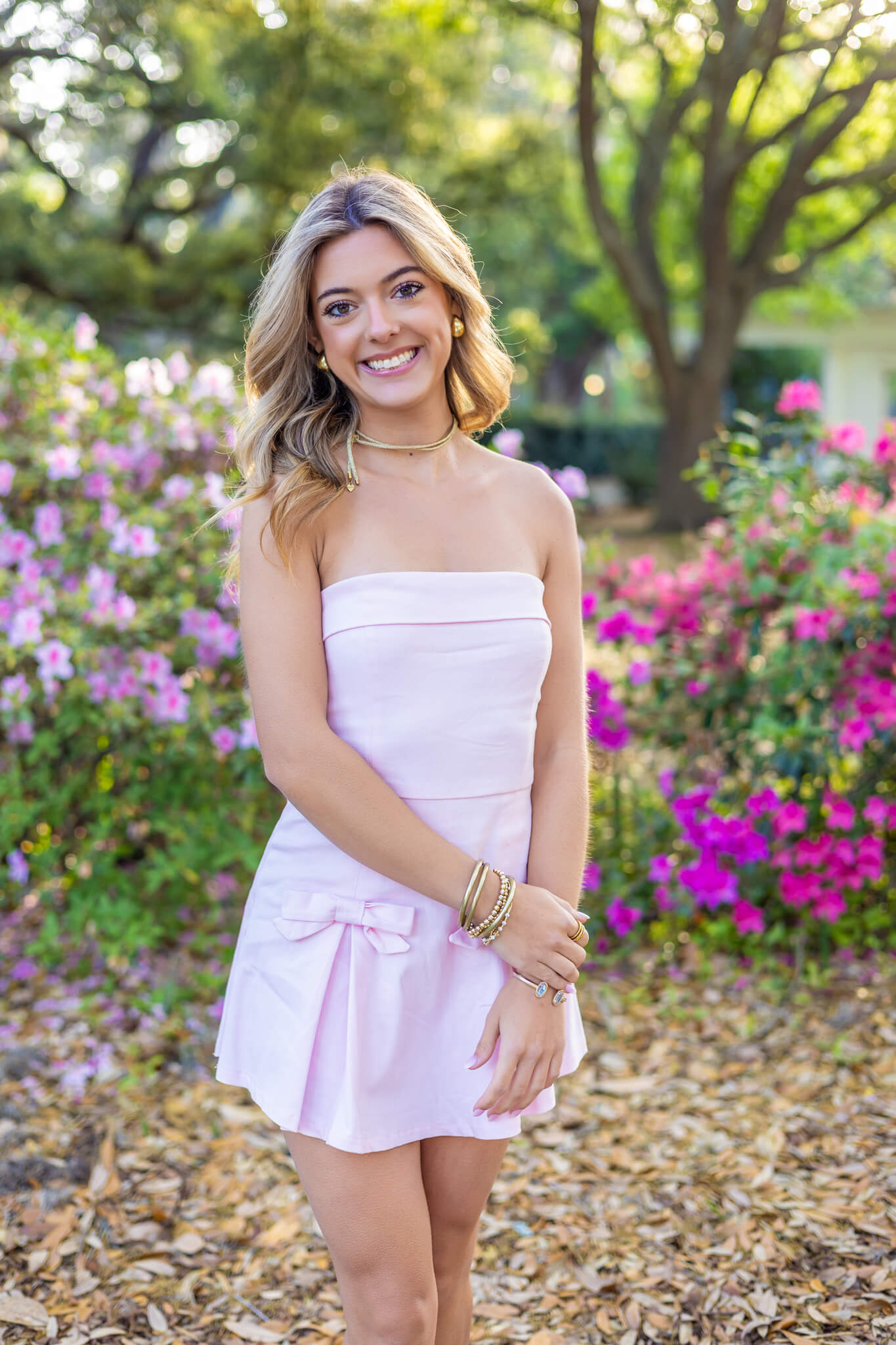 A smiling blonde woman in a pink dress stands in an azalea garden at sunset after shopping for prom dresses in Pooler, GA
