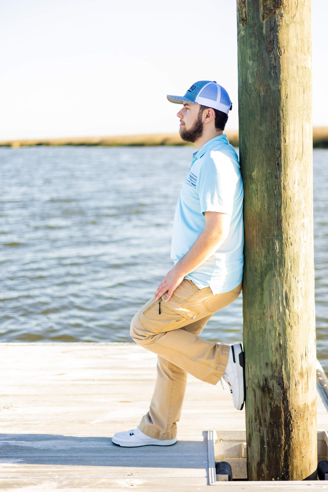 A happy high school senior leans on a wooden dock in a blue and white hat and blue polo