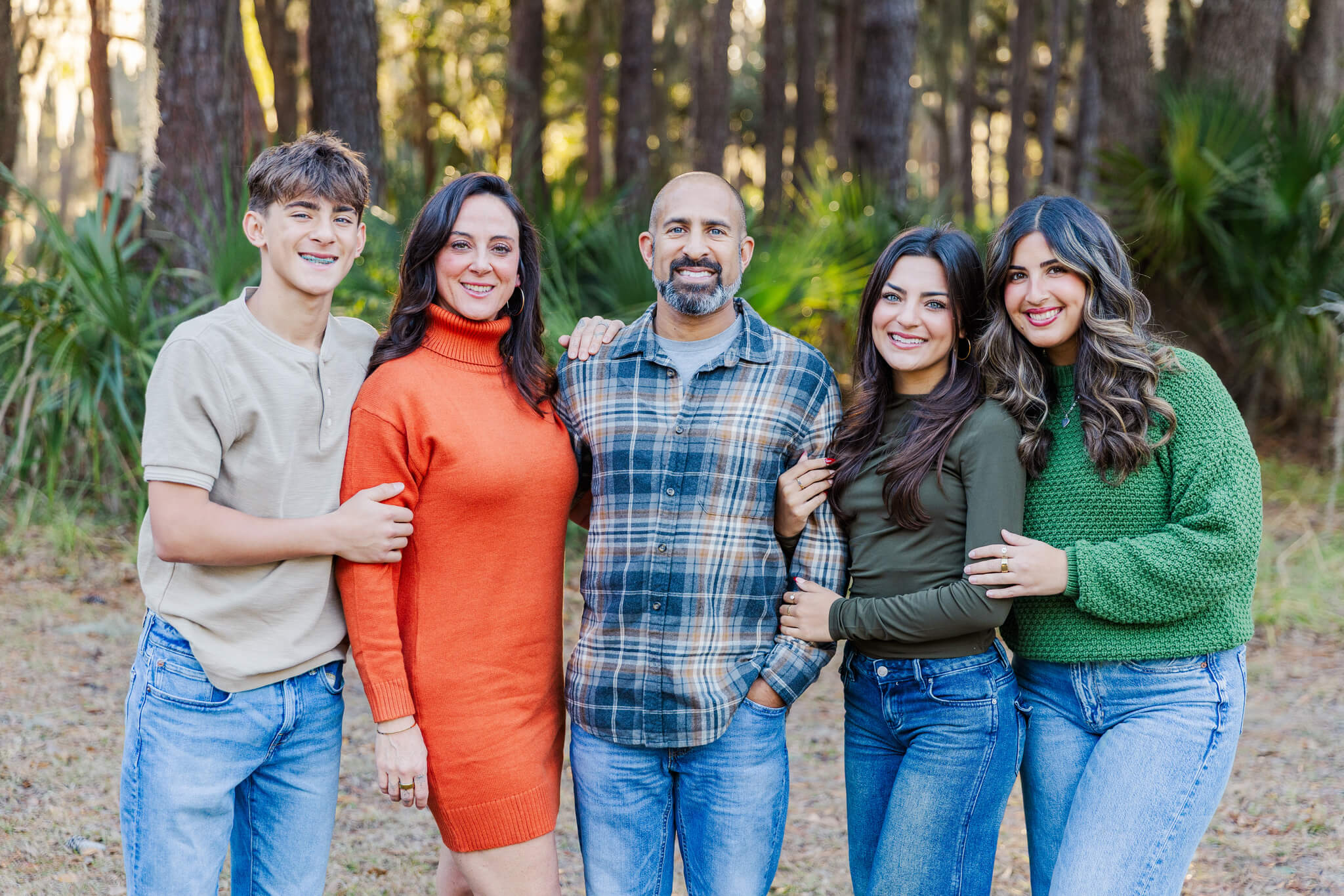 A smiling mom and dad stand in a park on the edge of a forest with their three teen children hugging onto them