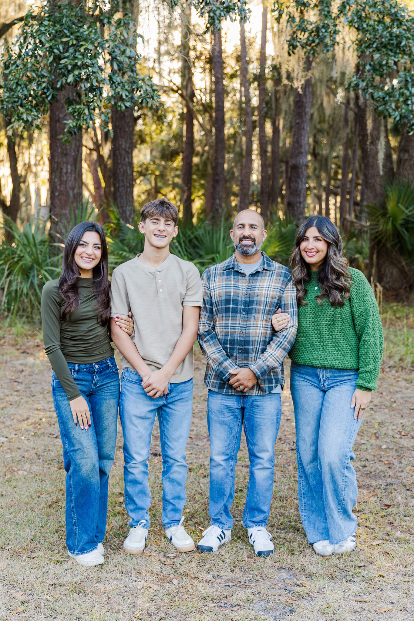A proud dad stands with his teen son and two daughters in a park smiling in jeans and green tops after enjoying date night in Pooler, GA