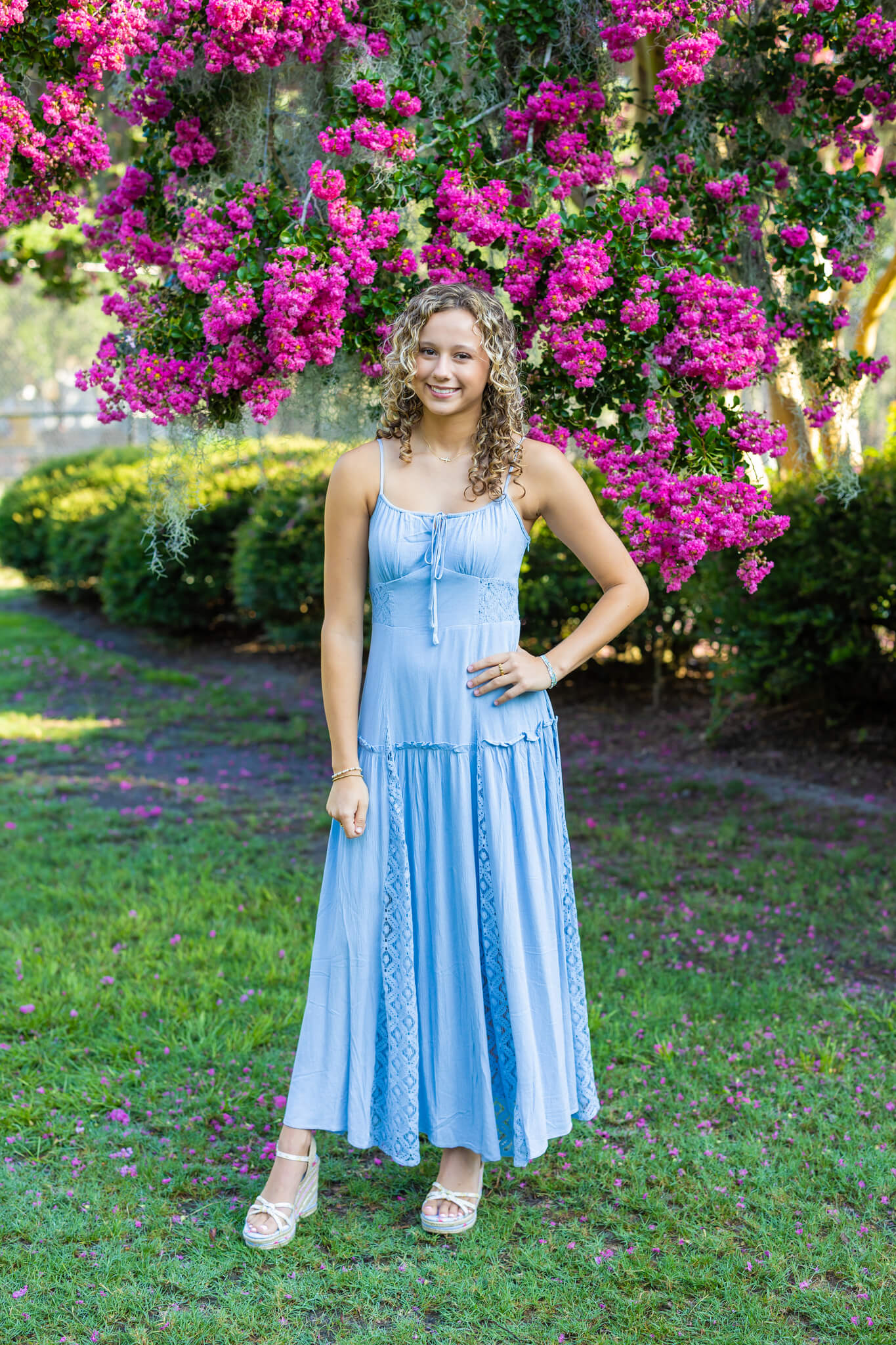 A smiling high school senior in a blue dress and heals stands with a hand on her hip under a pink tree in a park