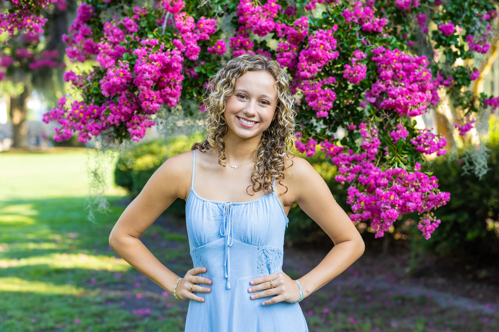 A happy high school senior in a blue dress smiles with hands on her hips under a pink blooming tree at one of the event spaces in pooler, ga