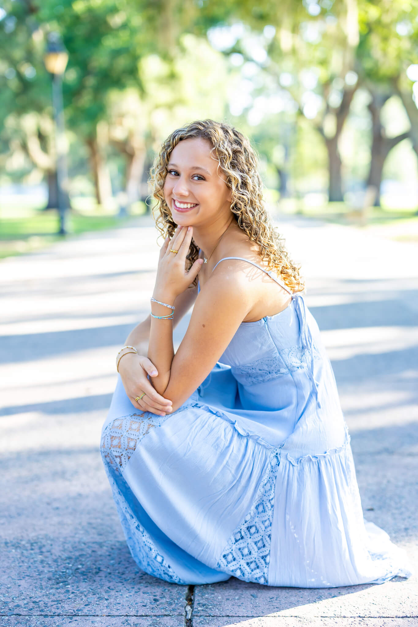 A smiling high school senior kneels in a sidewalk in a blue dress after visiting event spaces in pooler, ga