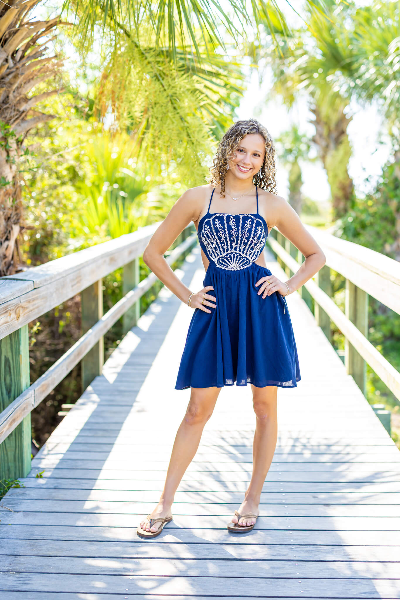 A proud high school grad leans in a blue dress with hands on her hips on a beach boardwalk after visiting hair salons in pooler ga