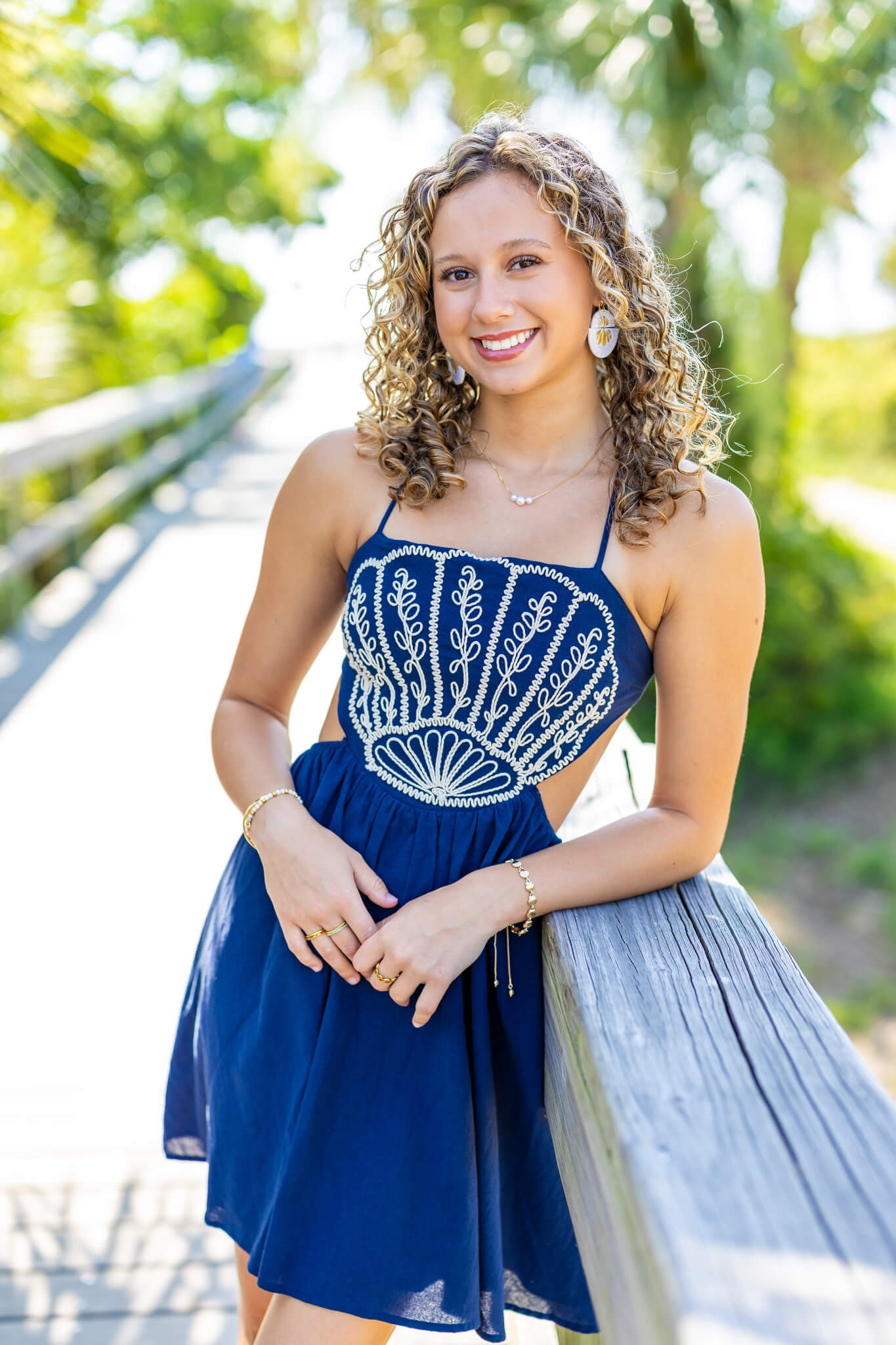 A smiling curly hair high school senior in a blue dress leans on a wooden park bridge after visiting hair salons in pooler ga