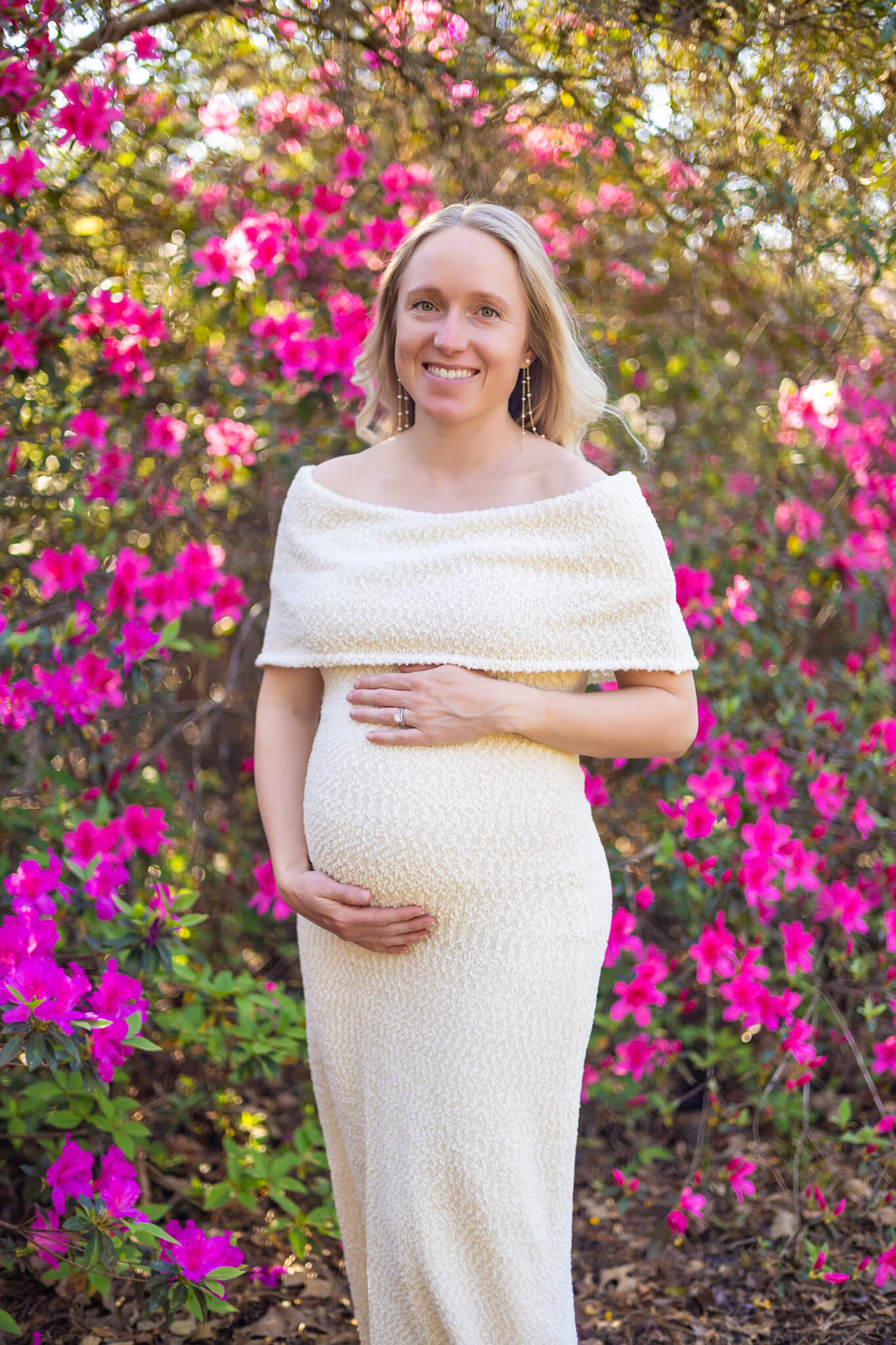 A smiling mother to be stands in a white maternity gown in an azalea garden