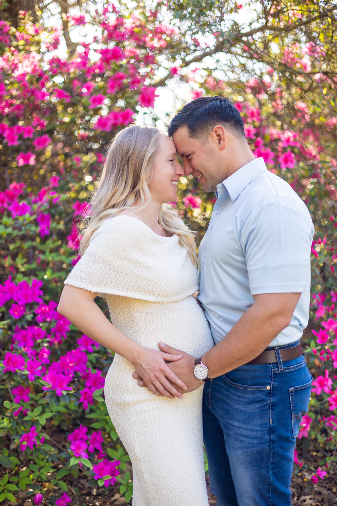 A happy expectant couple stands touching foreheads in white and blue in an azalea garden after finding perfect jewelry stores in savannah, ga