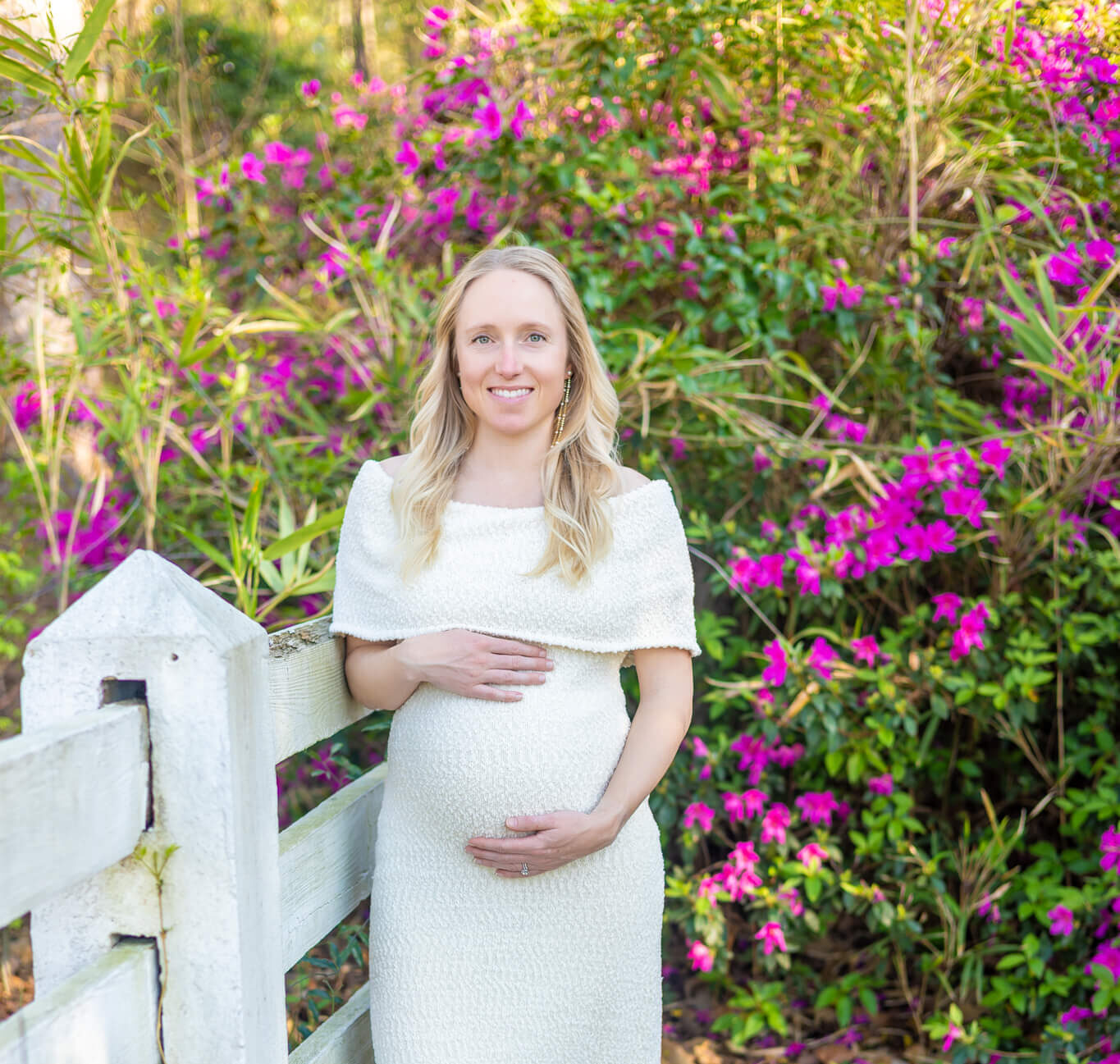 A pregnant woman stands against a rustic garden fence holding her bump in a white dress after visiting jewelry stores in savannah, ga