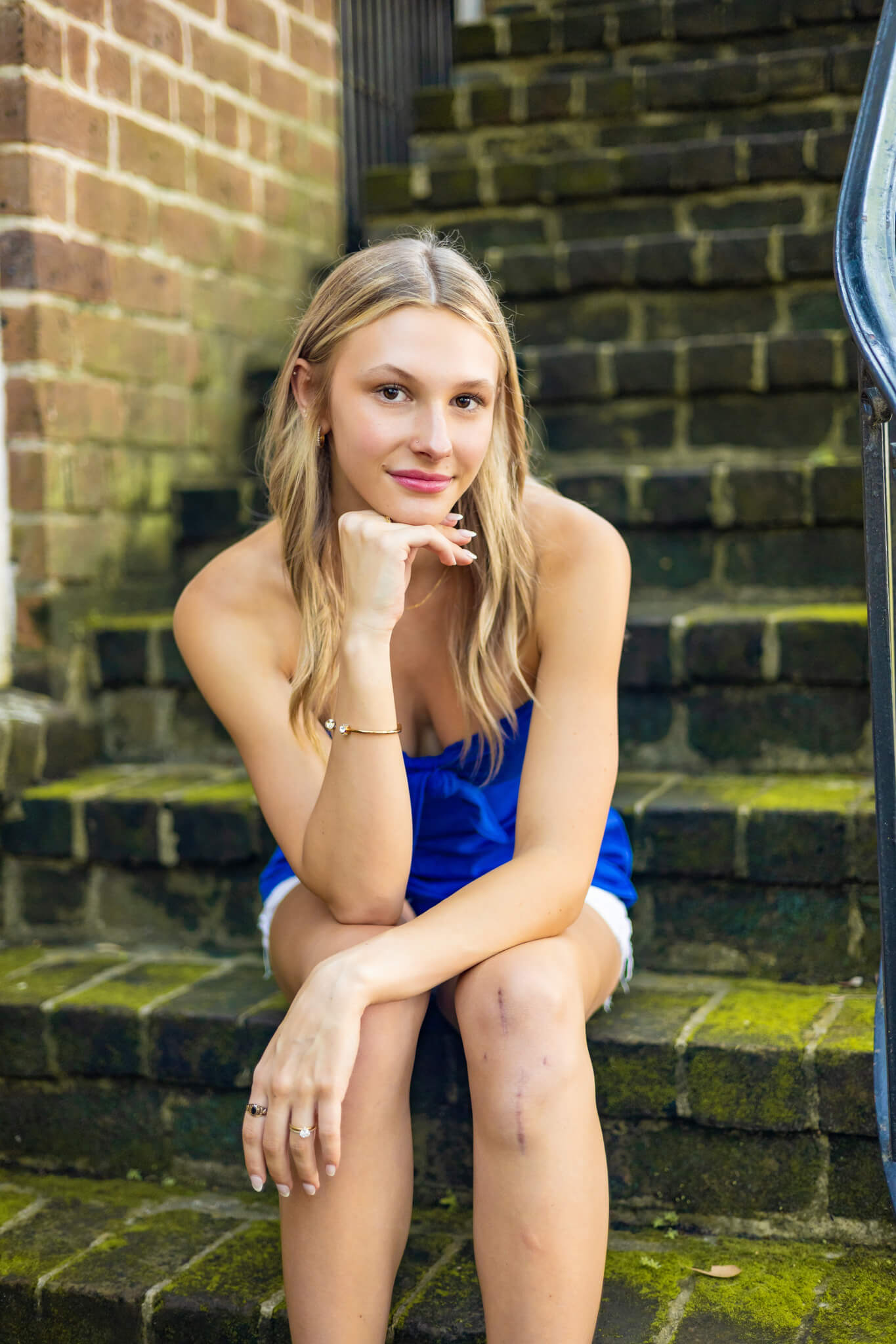 A woman leans on her kneels while sitting on rustic brick stairs in a blue blouse