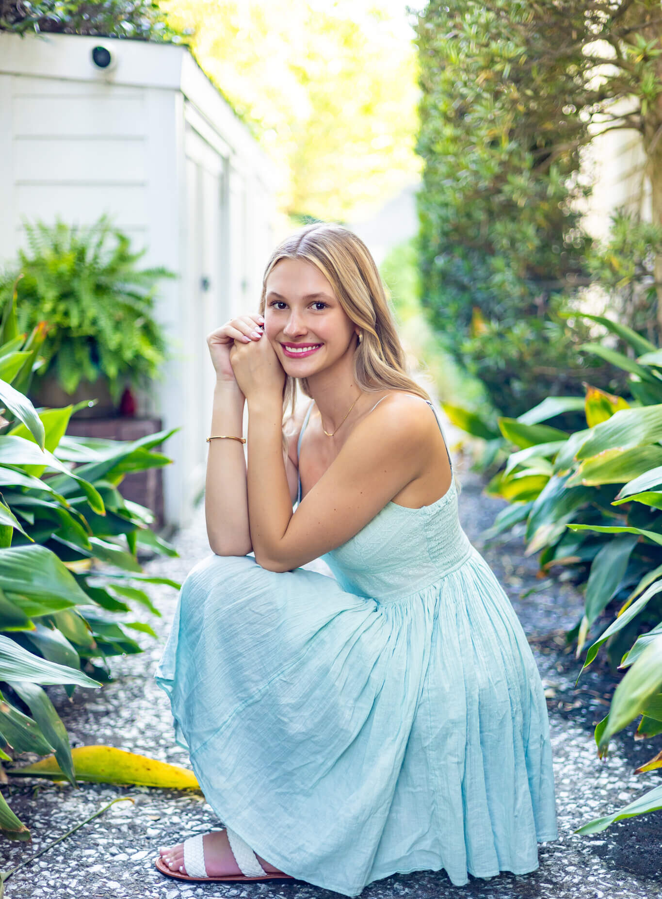 A smiling woman in a blue dress kneels in a garden path smiling after finding nail salons in pooler ga