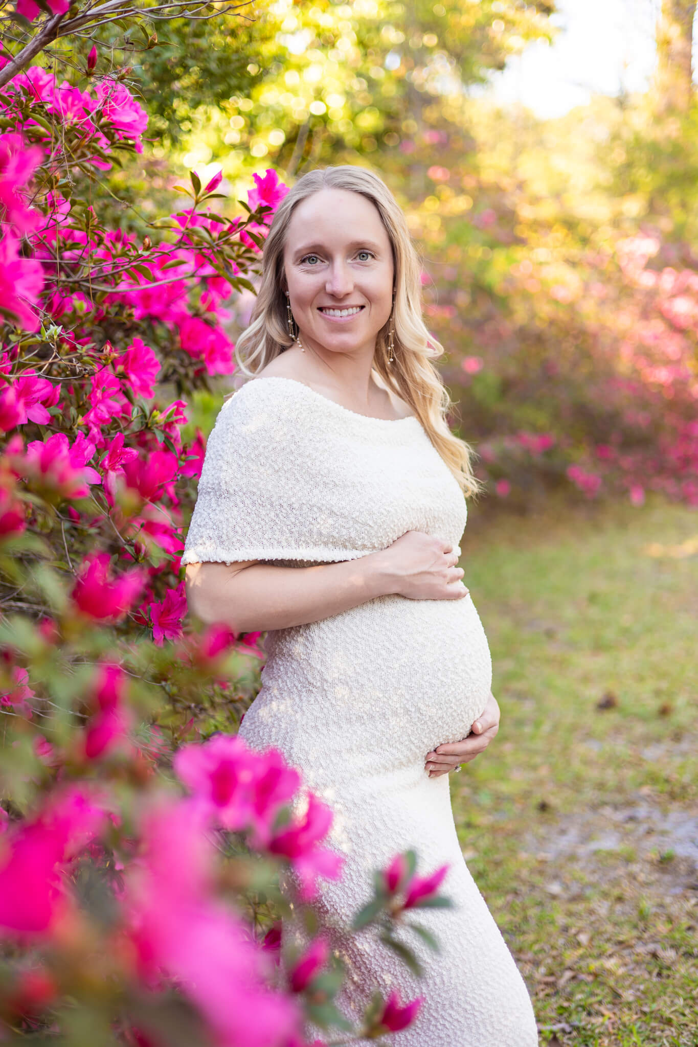 A smiling pregnant woman stands in an azalea garden in a white maternity dress at sunset with hands on the bump