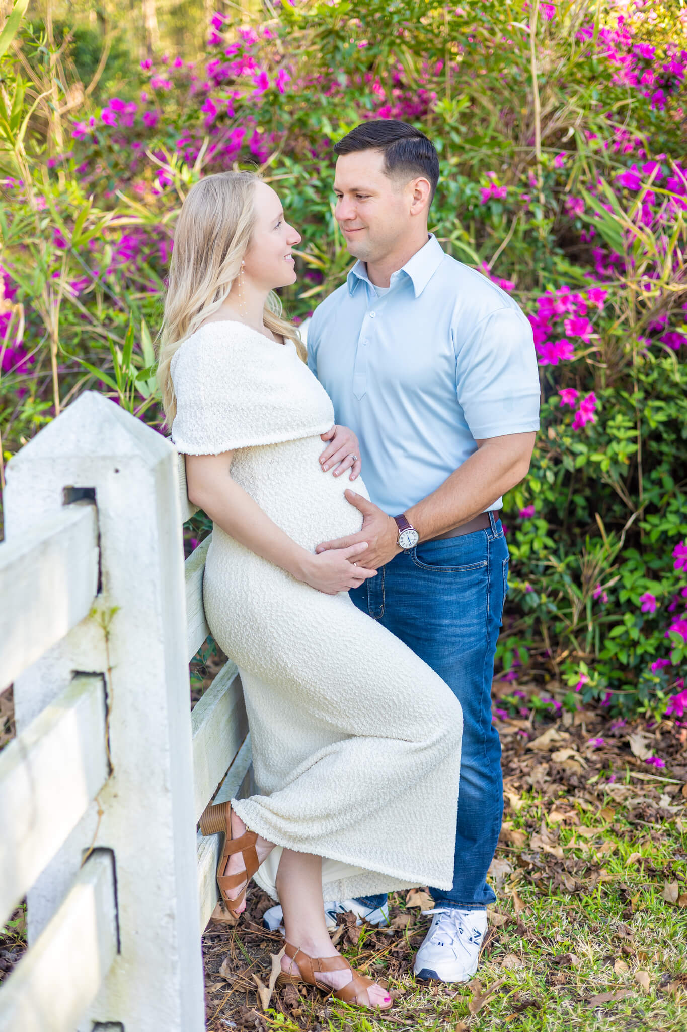 A happy couple lean against a garden fence in white and blue outfits with hands on the pregnant belly after he was able to propose in savannah