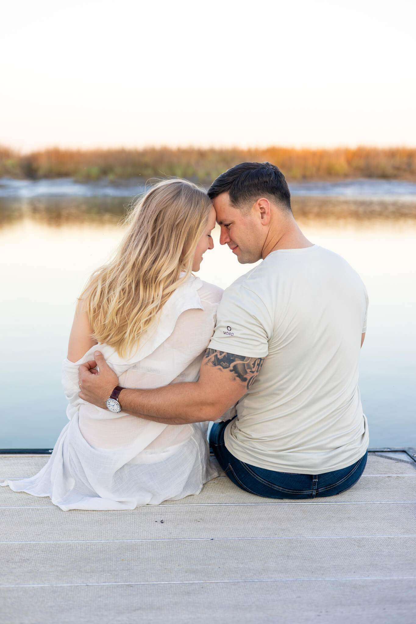 A happy couple in white snuggle while touching foreheads and sitting on the edge of a dock after planning to propose in savannah
