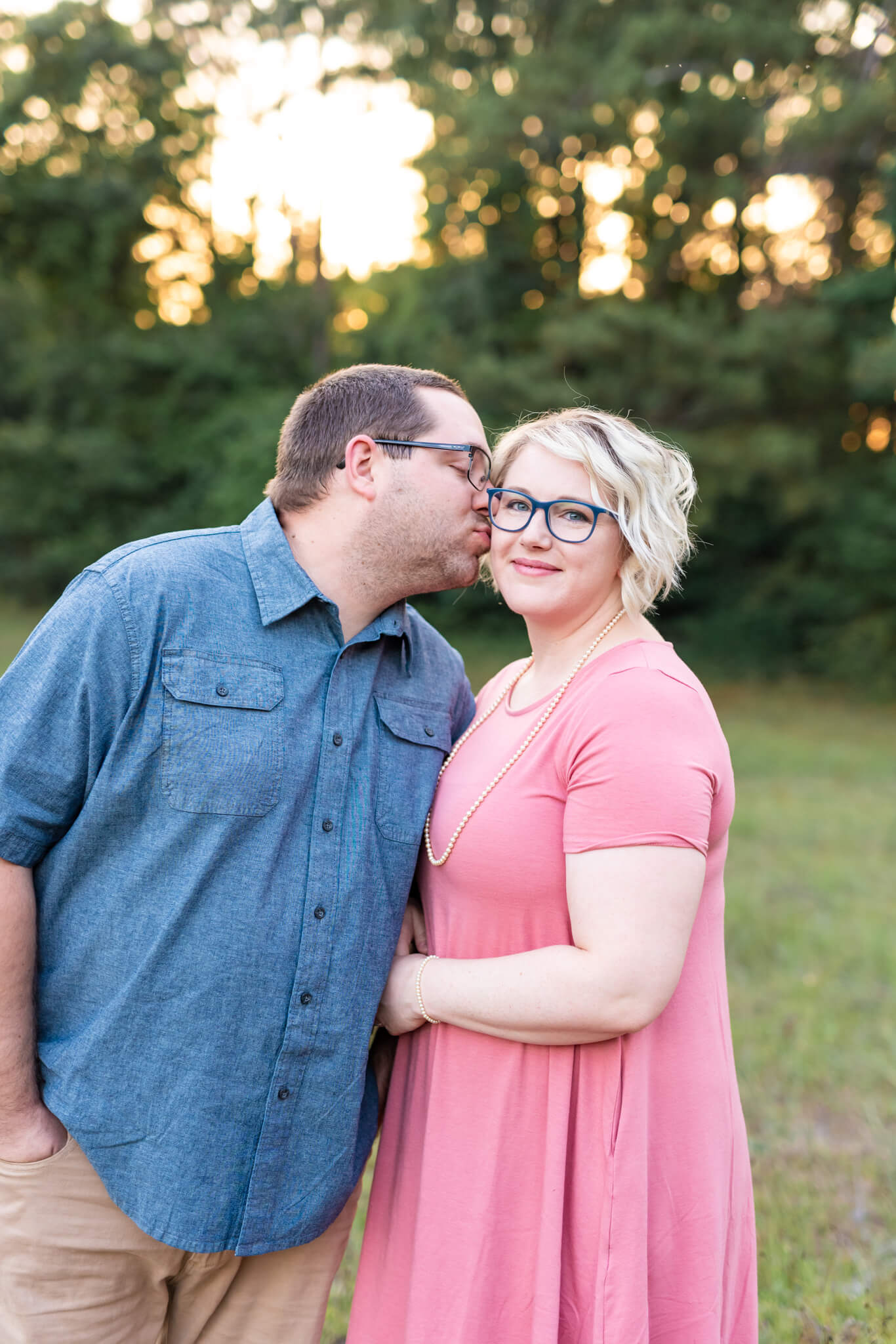 A happy woman in a pink dress is kissed by her husband in blue in a park lawn at sunset after enjoying romantic restaurants in Pooler, GA