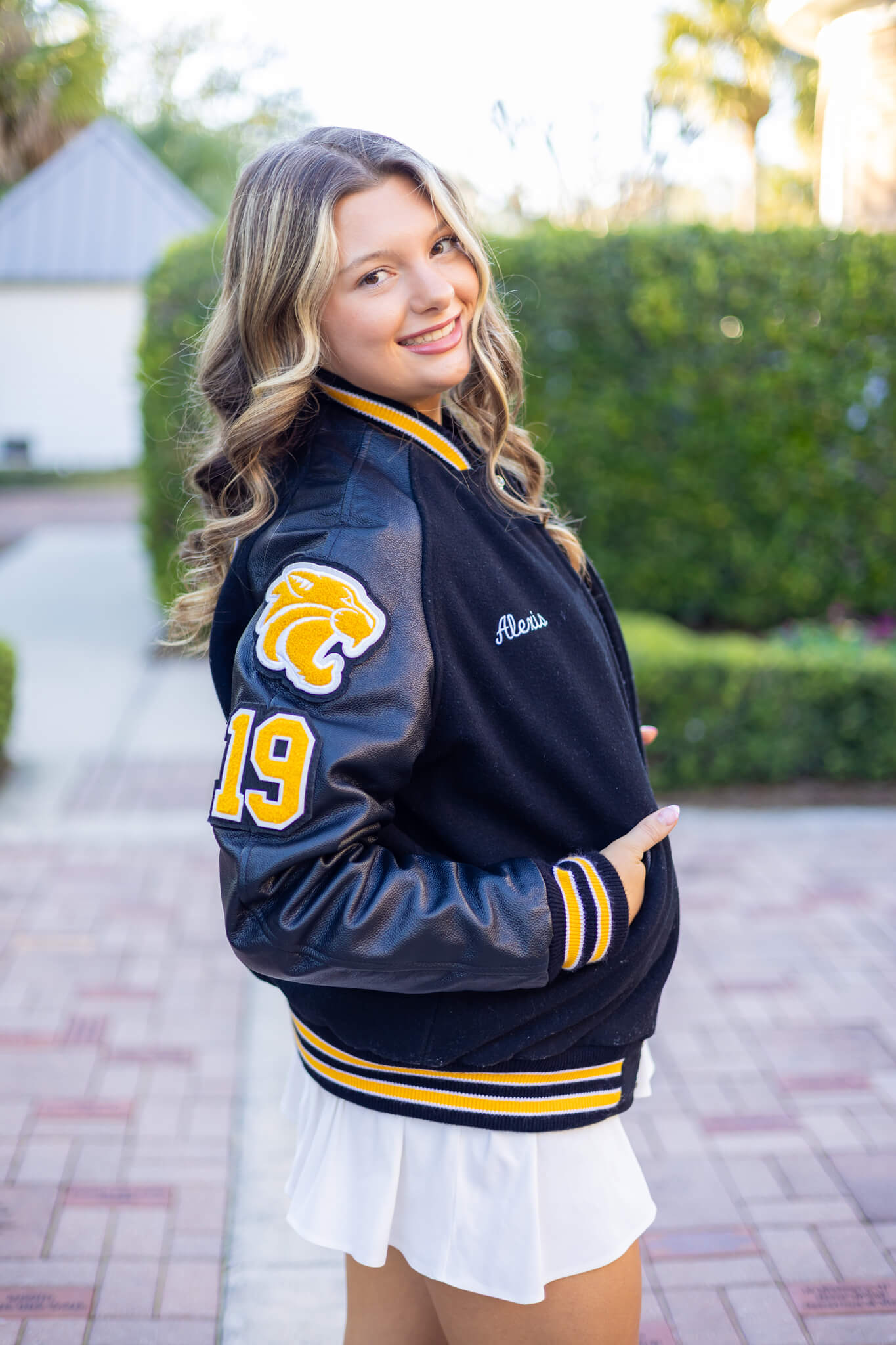 A happy high school senior in a black letterman jacket and white skirt walks in a garden smiling over her shoulder after visiting tanning salons in pooler ga