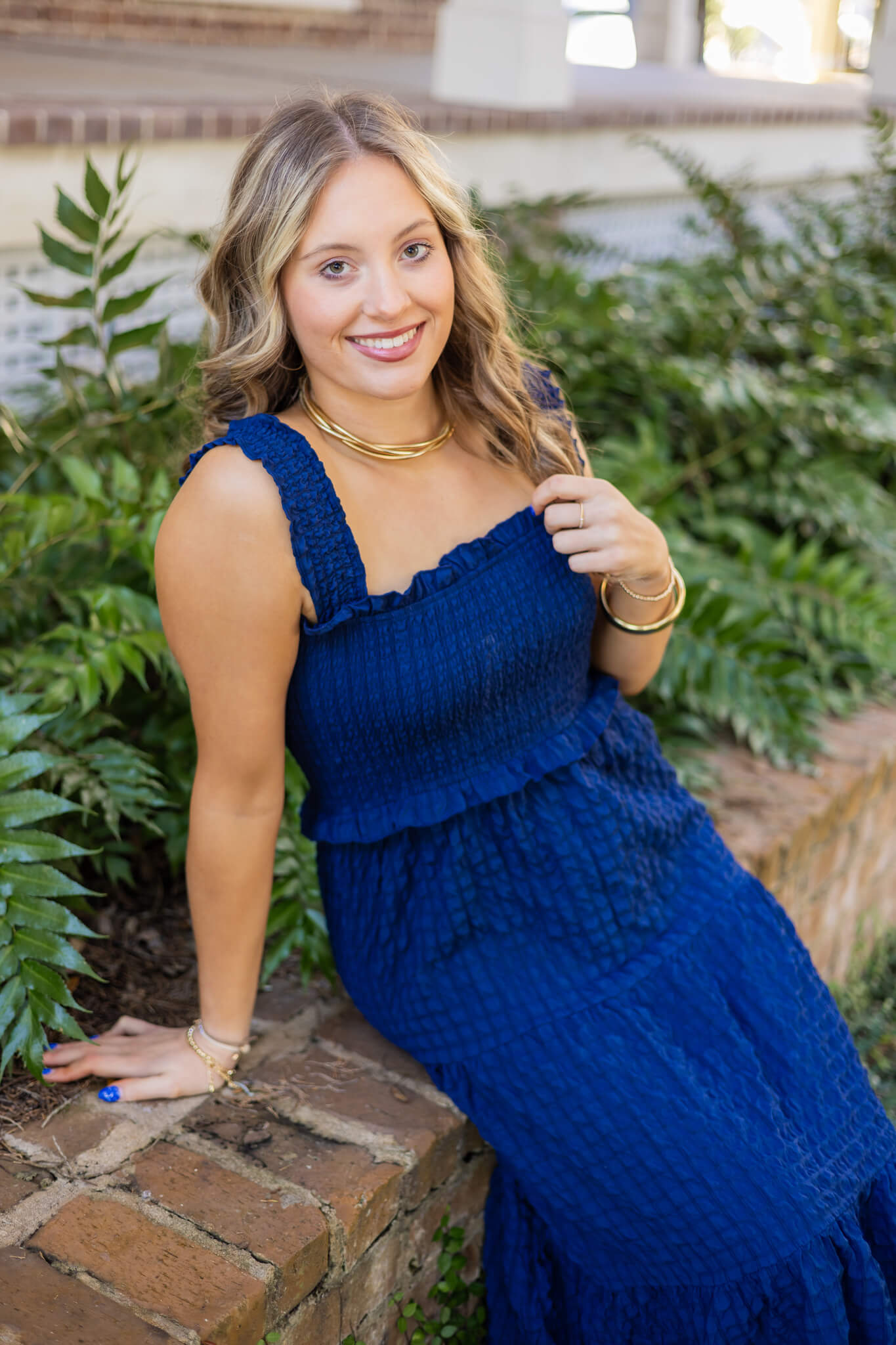 A high school senior in a blue dress sits on a garden brick wall smiling after finding tanning salons in pooler ga