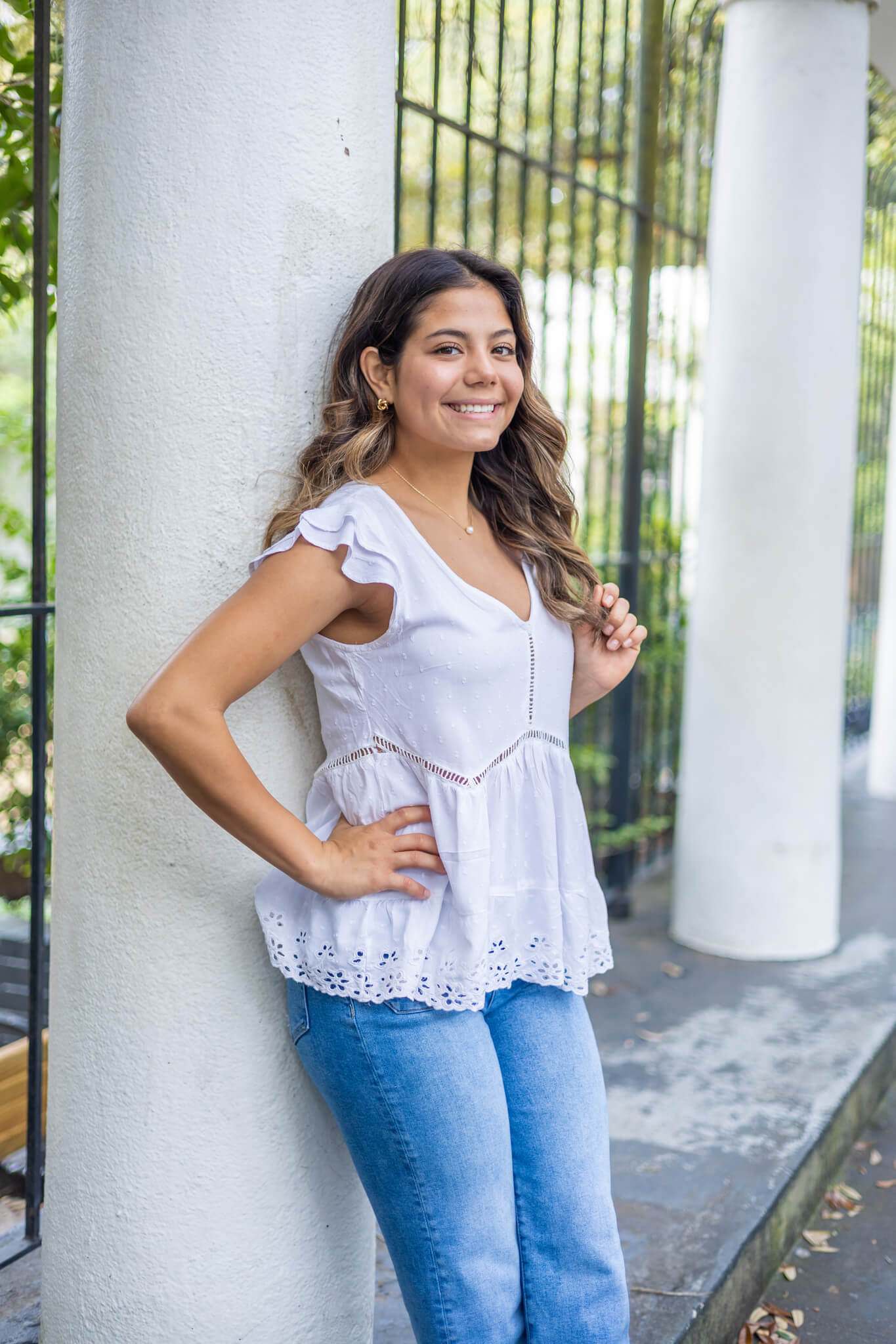 A happy high school senior in a white blouse and jeans leans on a park column twirling her hair after enjoying boutiques in Richmond Hill, GA
