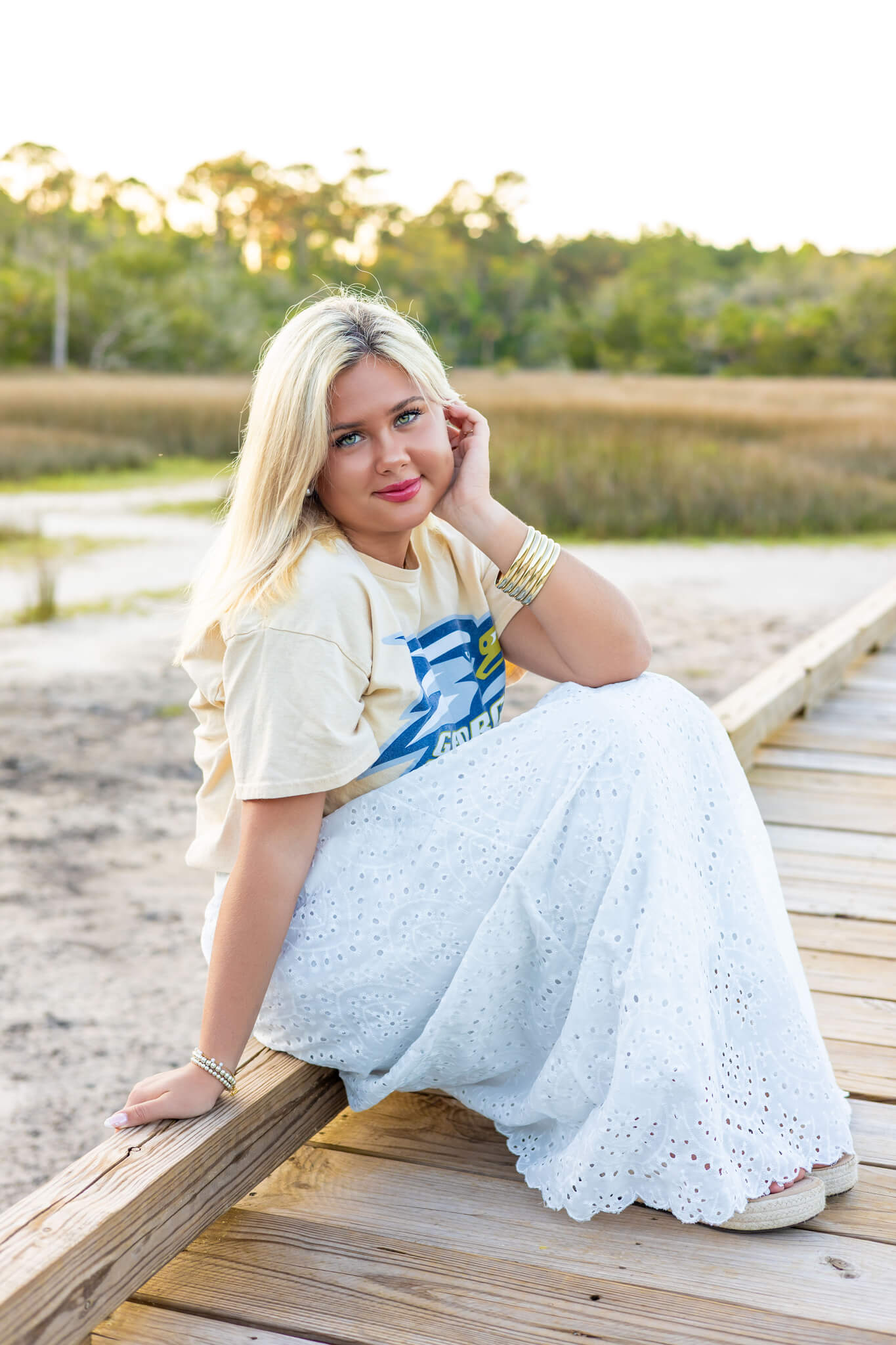 A happ high school senior sits on a beach boardwalk at sunset pulling hair behind her ear