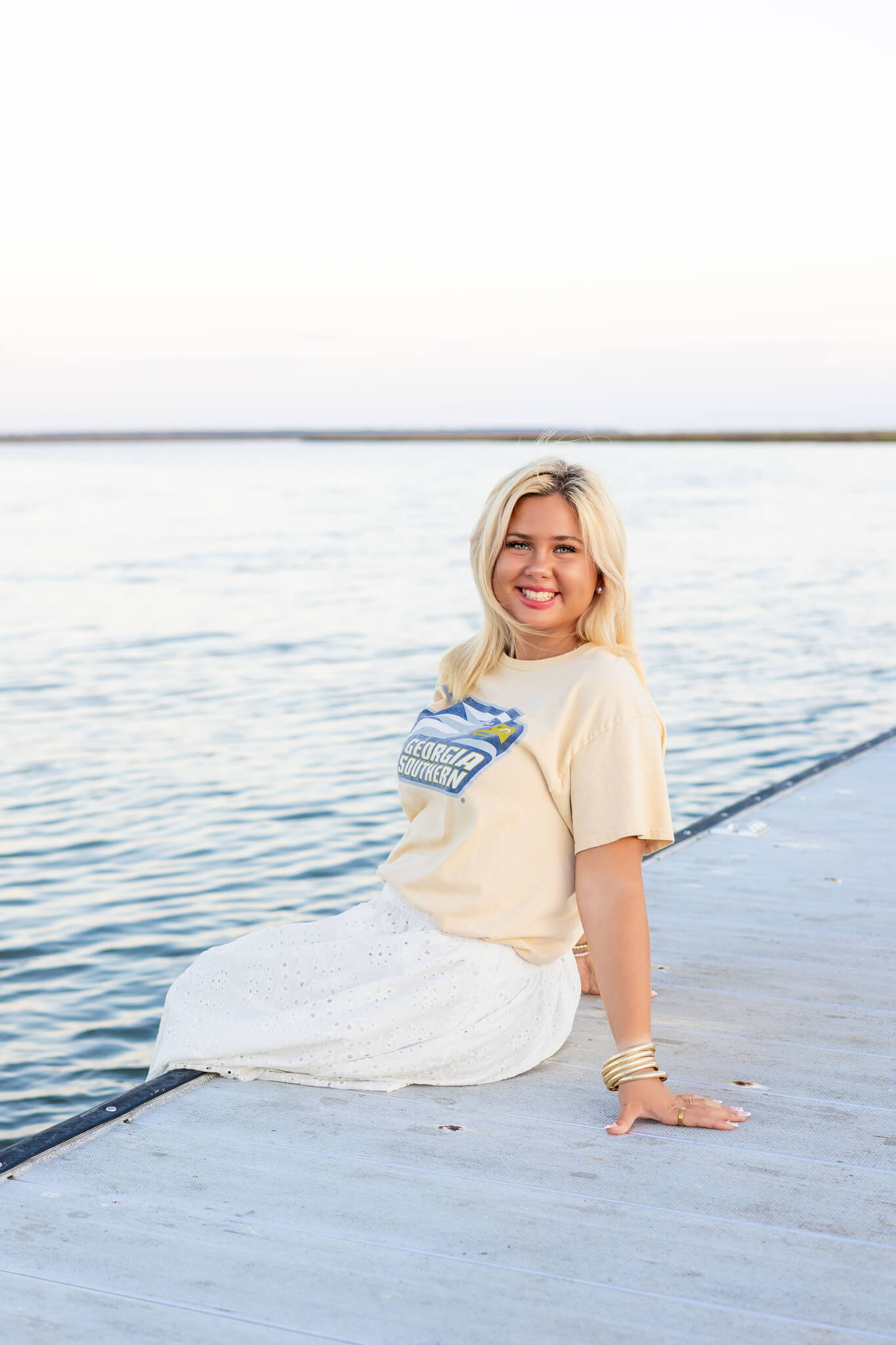 A smiling high school senior in a yellow shirt sits on a dock smiling big at sunset after visiting colleges near pooler ga