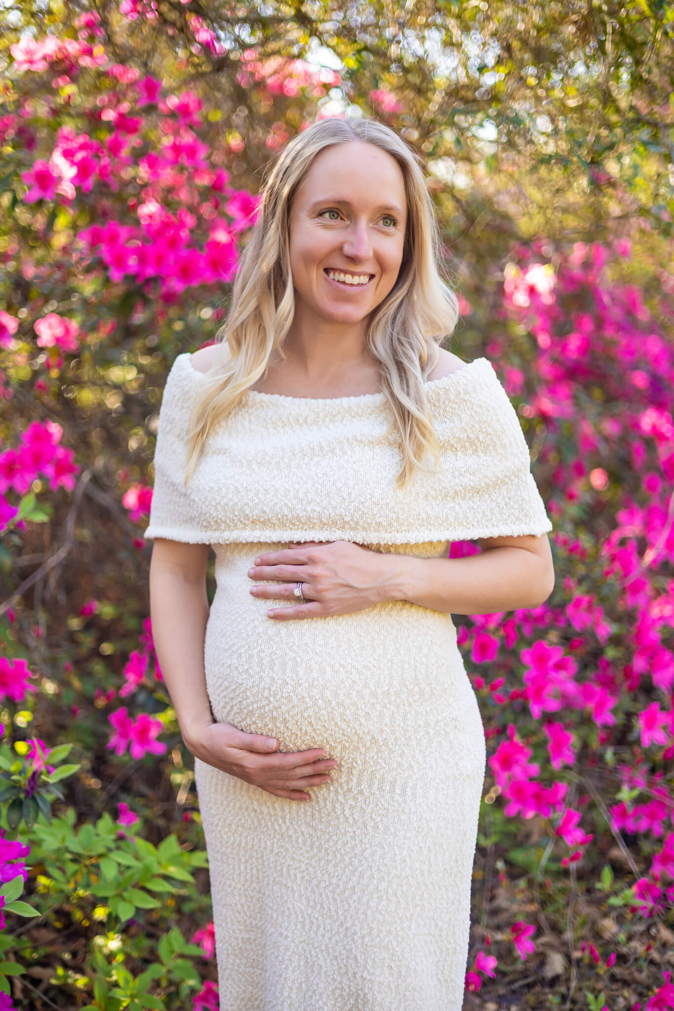 A happy pregnant woman smiles while standing in front of pink azalea bush