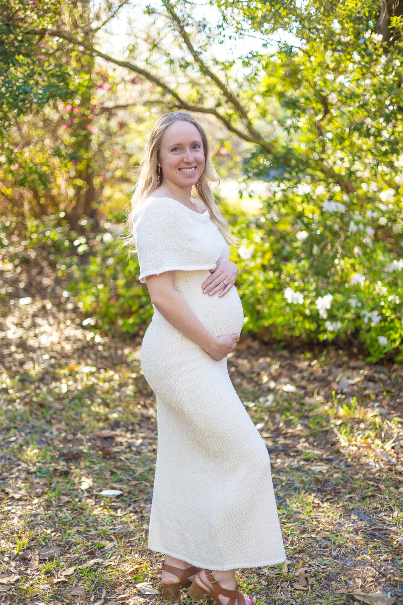 A mom to be stands holding her bump in a white dress in a park at sunset after enjoying a couples massage in Pooler, GA