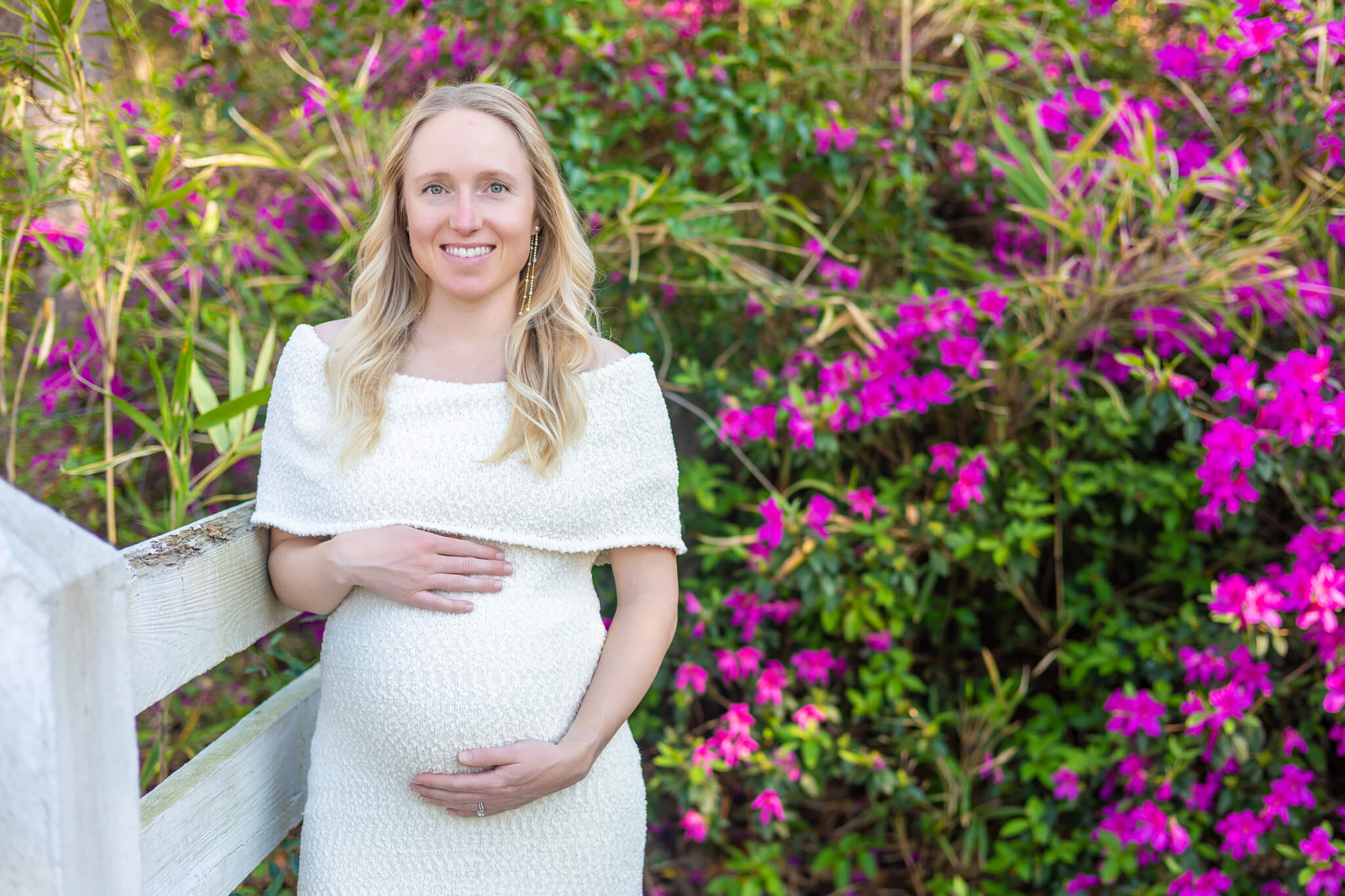 A pregnant woman leans against a white picket fence in an azalea garden at sunset in a white maternity dress after enjoying a couples massage in Pooler, GA