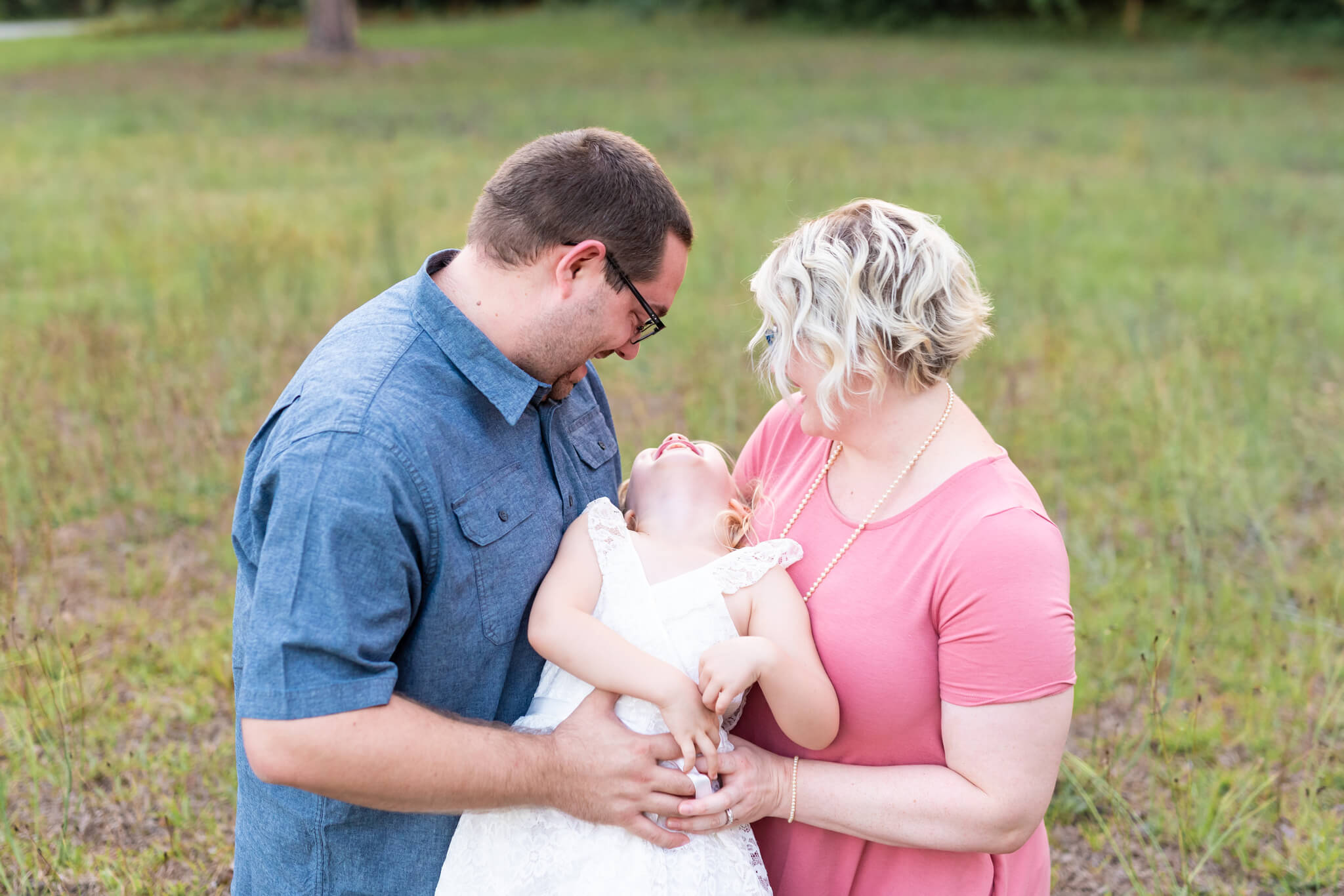 A toddler girl in a white dress laughs while being tickled by mom and dad in a field