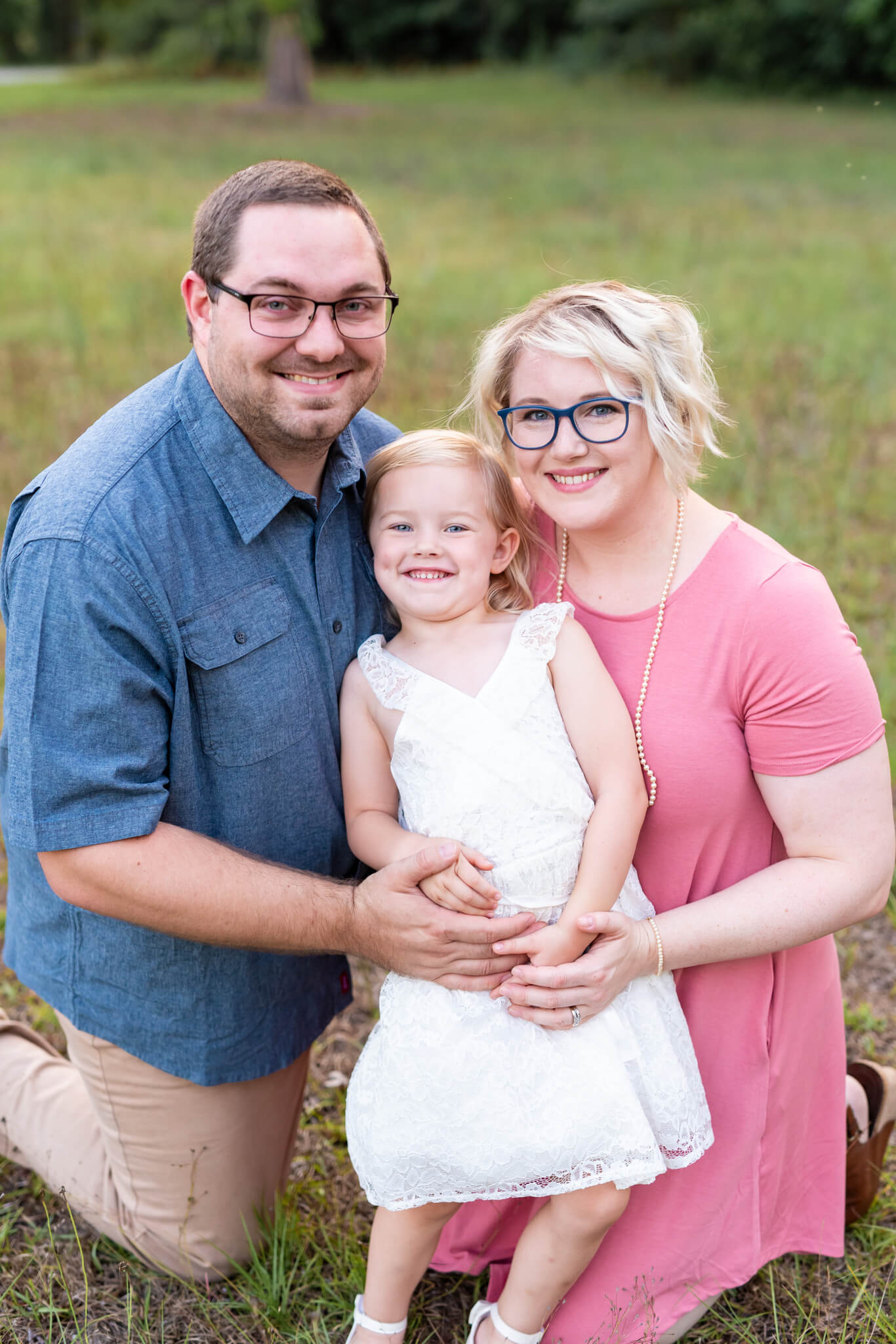 A mom and dad in blue and pink kneel in the grass with their giggling toddler daughter between them after finding date ideas in Richmond Hill, GA