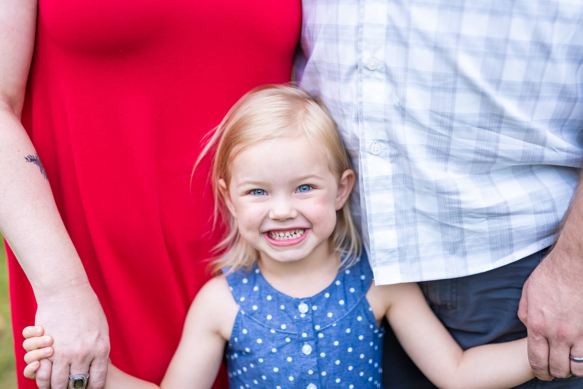 A happy toddler girl smiles while wearing a blue dress while holding mom and dad's hands