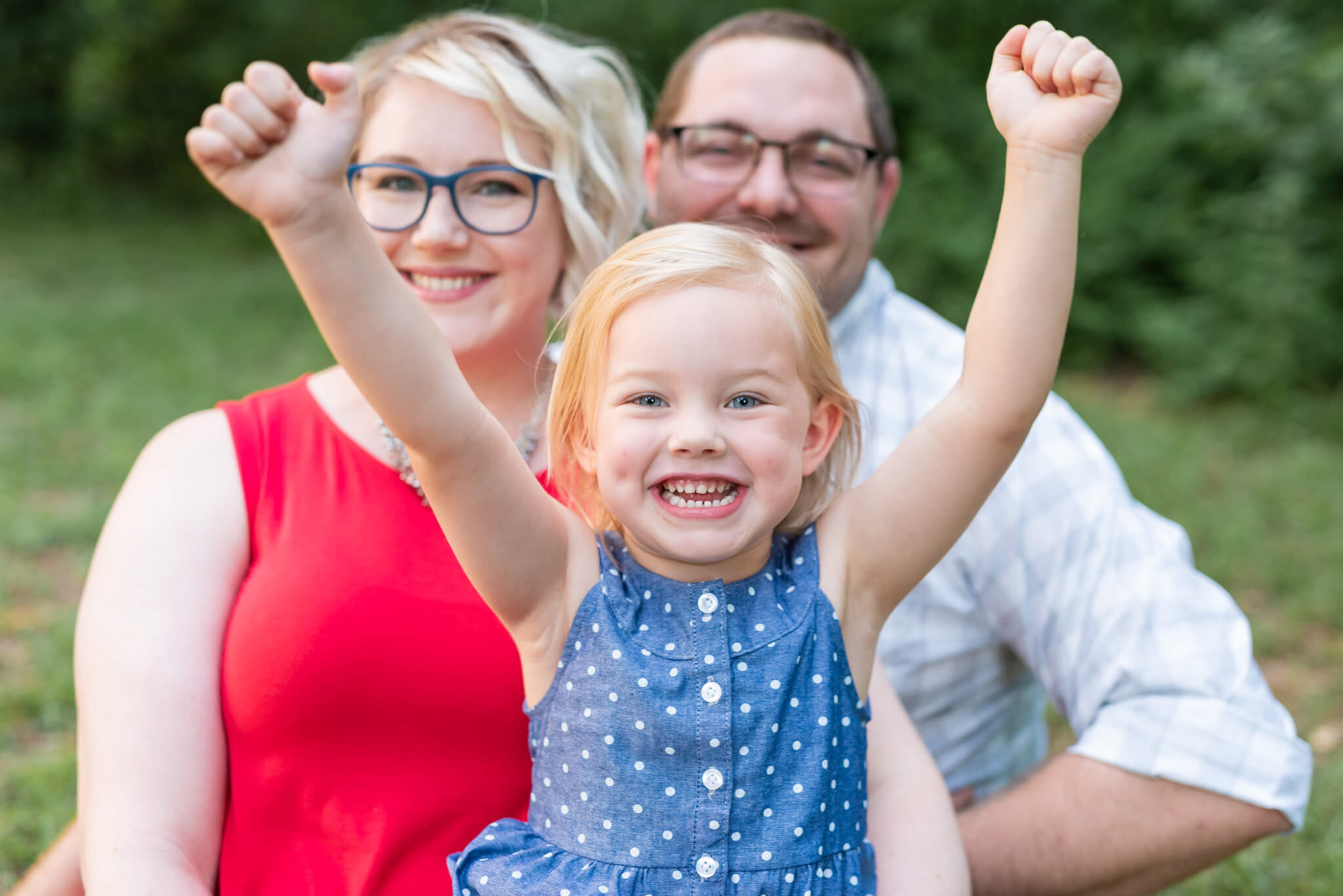 A mom and dad smile behind their cheering young daughter in a blue dress in front of them before visiting a day spa in pooler ga