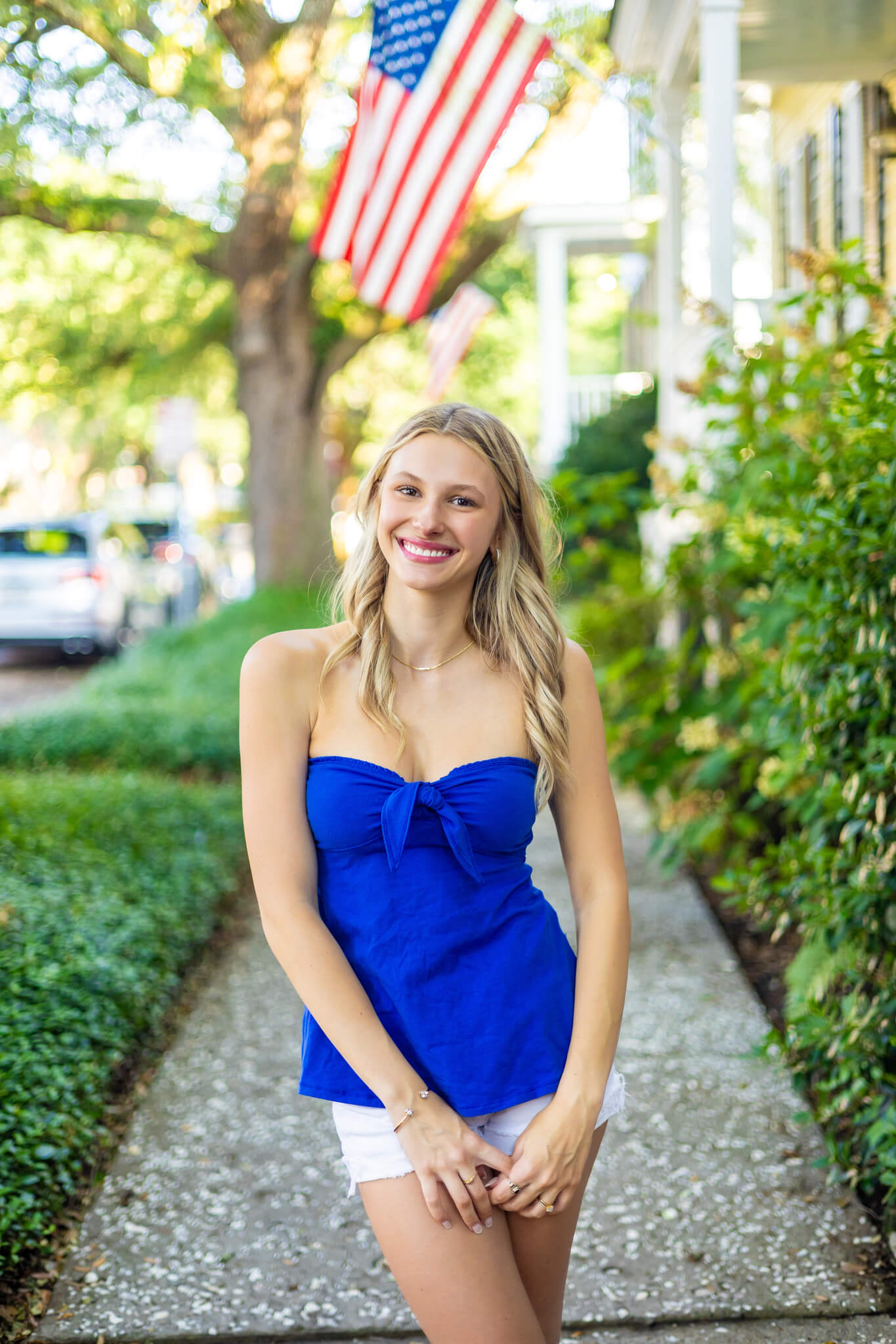 A smiling woman in a blue blouse and white shorts stands in a sidewalk