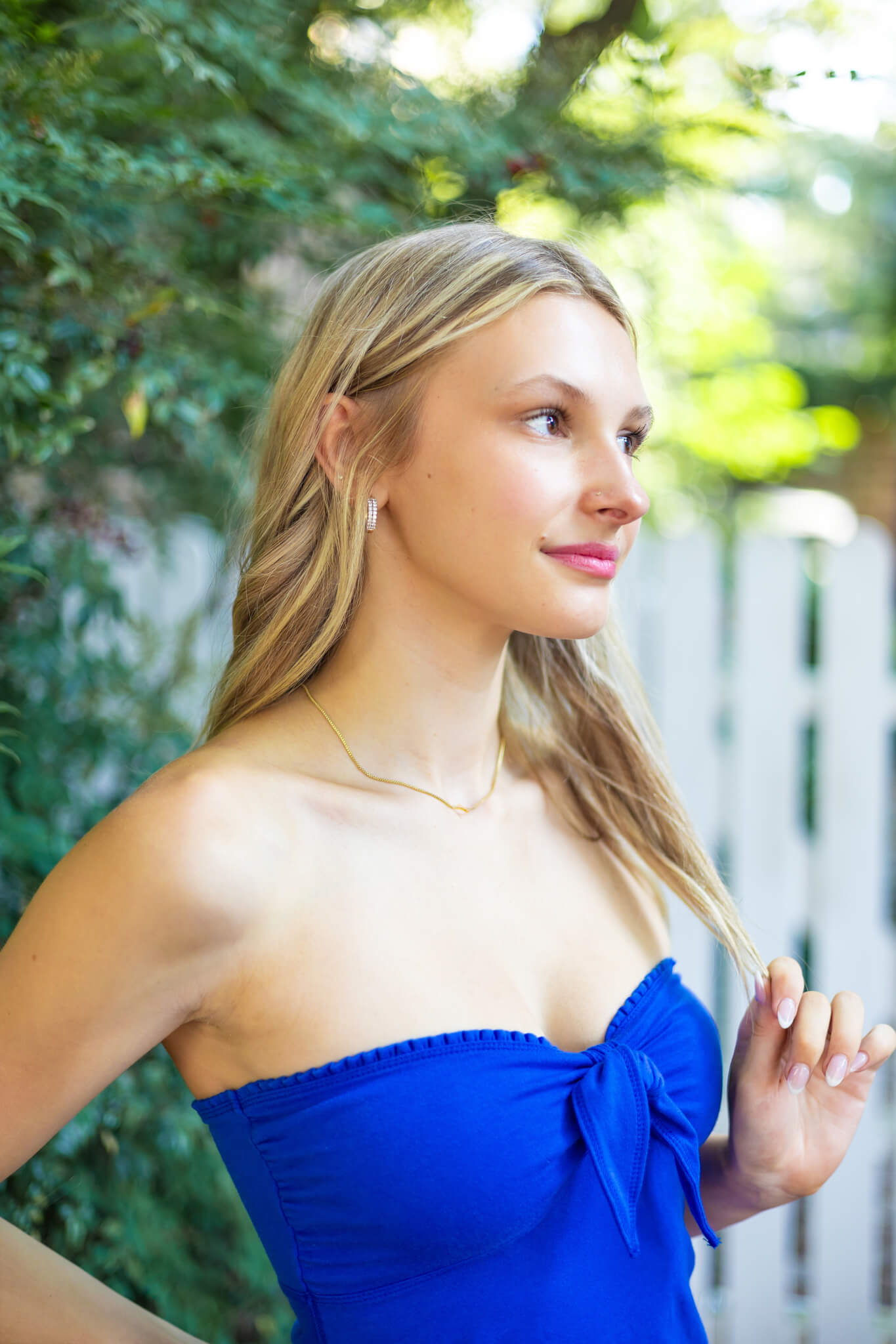 A blonde woman twirls her hair while standing by a white picket fence in a garden after visiting hair salons in Richmond Hill, GA in a blue blouse