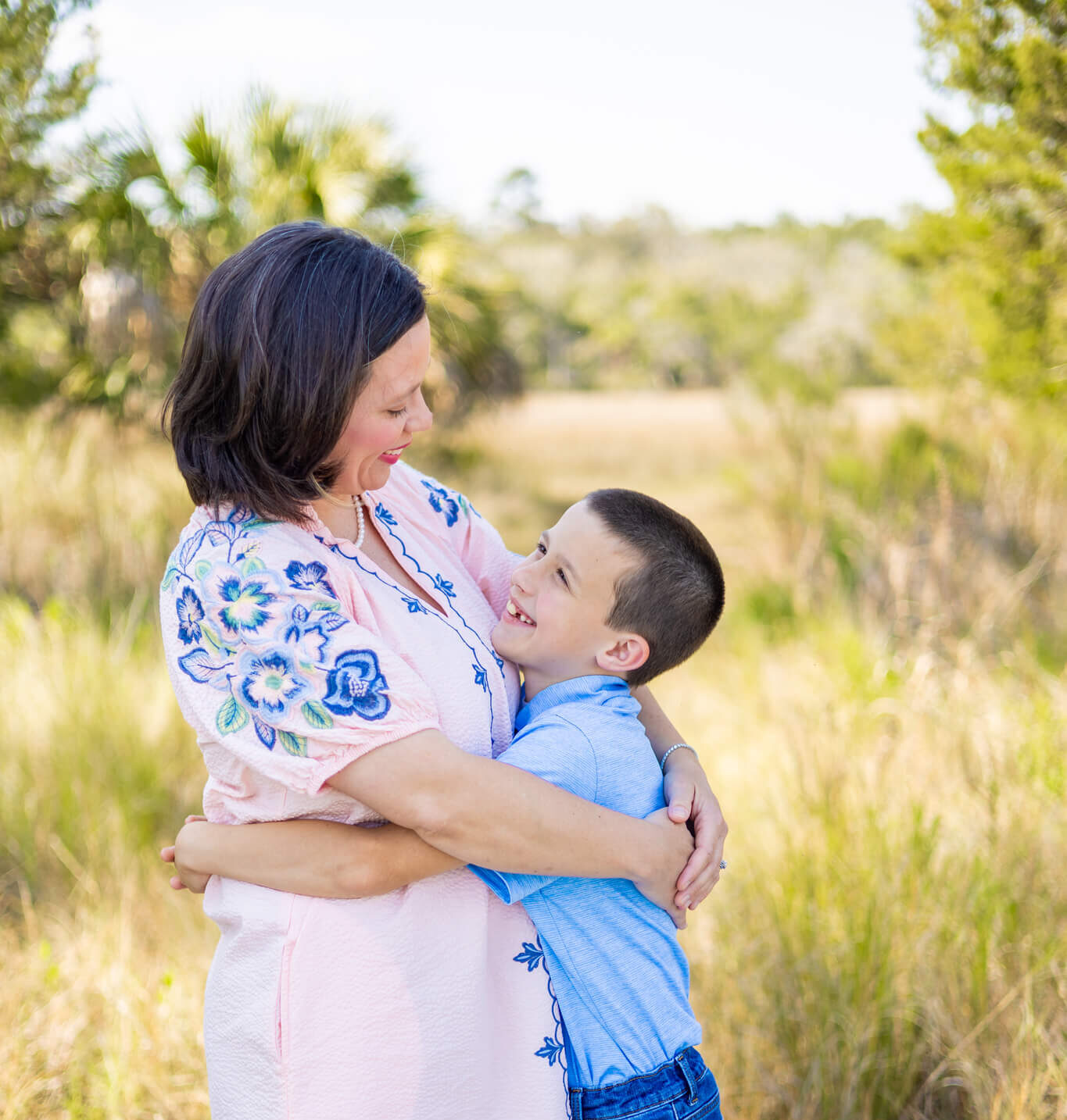 A happy young boy in a blue polo and jeans hugs mom in a pink flower print dress in a park field